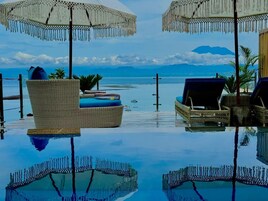 An infinity pool with lounge chairs and umbrellas, surrounded by a clear view of the sea and mountains.