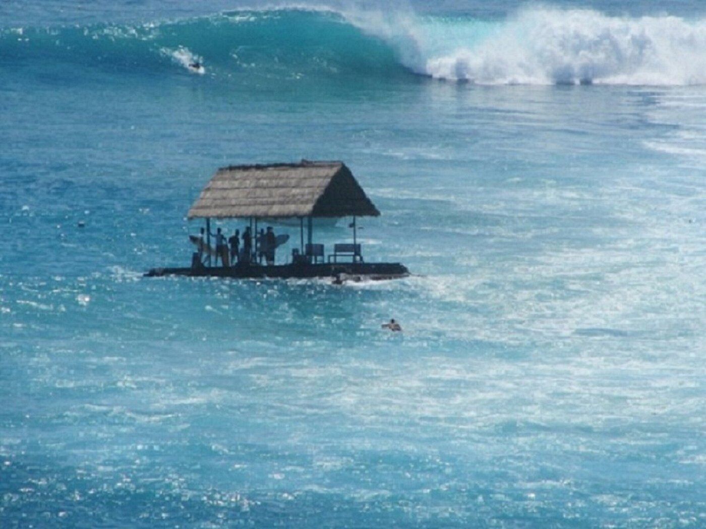A thatched-roof structure on a floating platform in the ocean, with people and a surfboard visible.