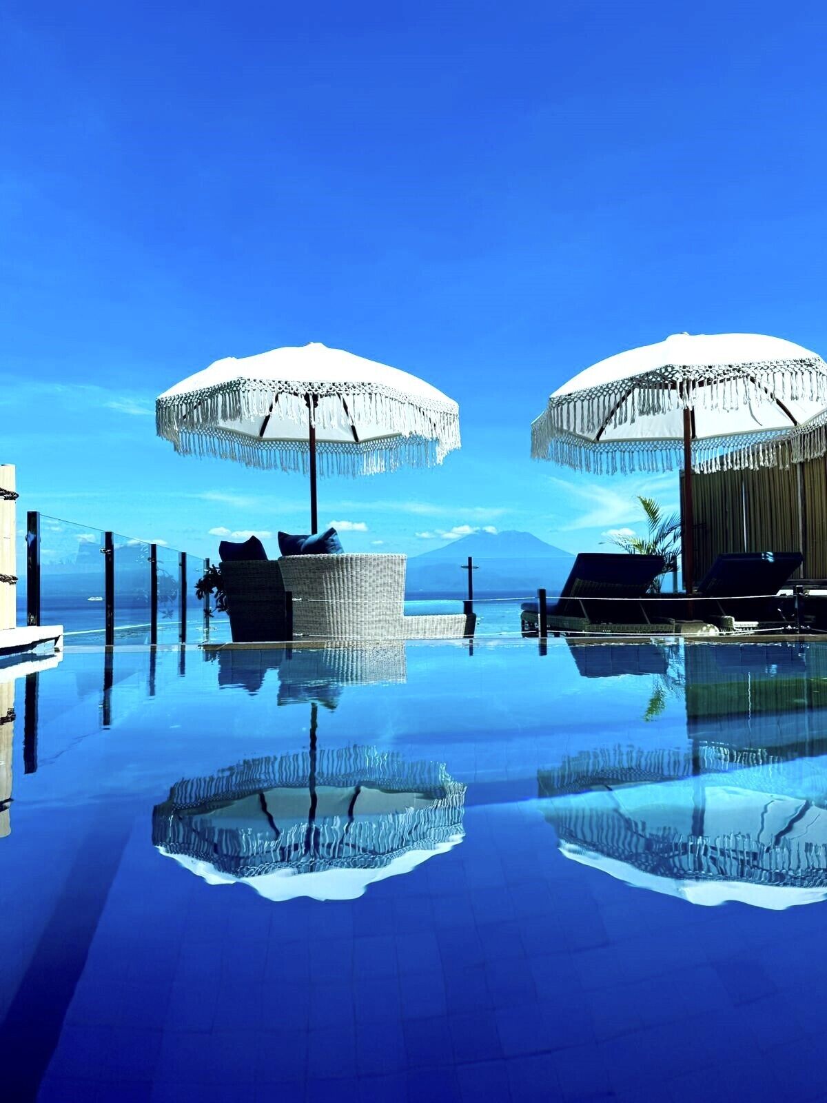 Ein Pool mit zwei weißen Sonnenschirmen und Liegestühlen, umgeben von klarem, blauen Wasser, das den Himmel spiegelt.