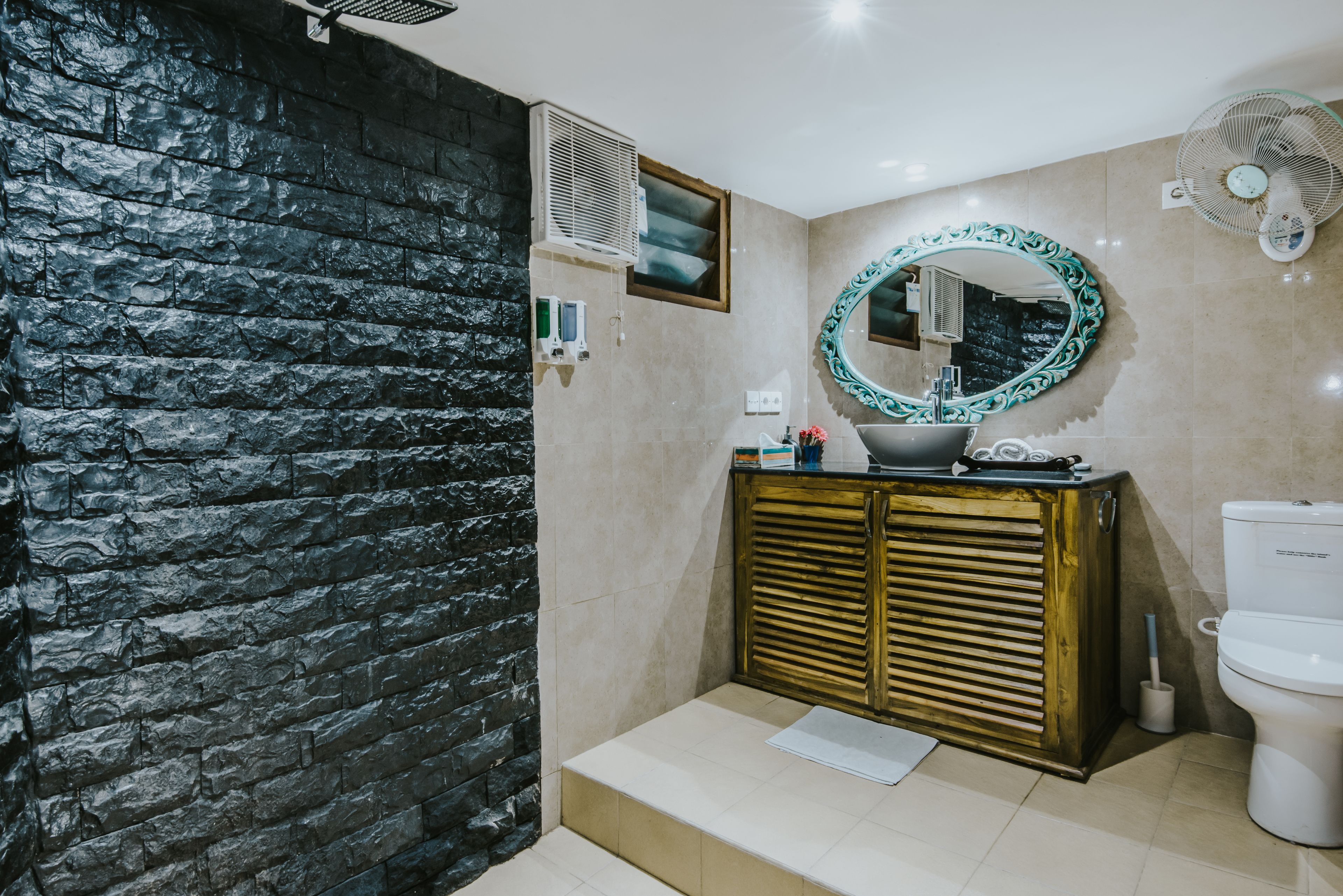 Een badkamer met een stenen muur, een wastafel met een decoratieve spiegel, een toilet en een ventilator.