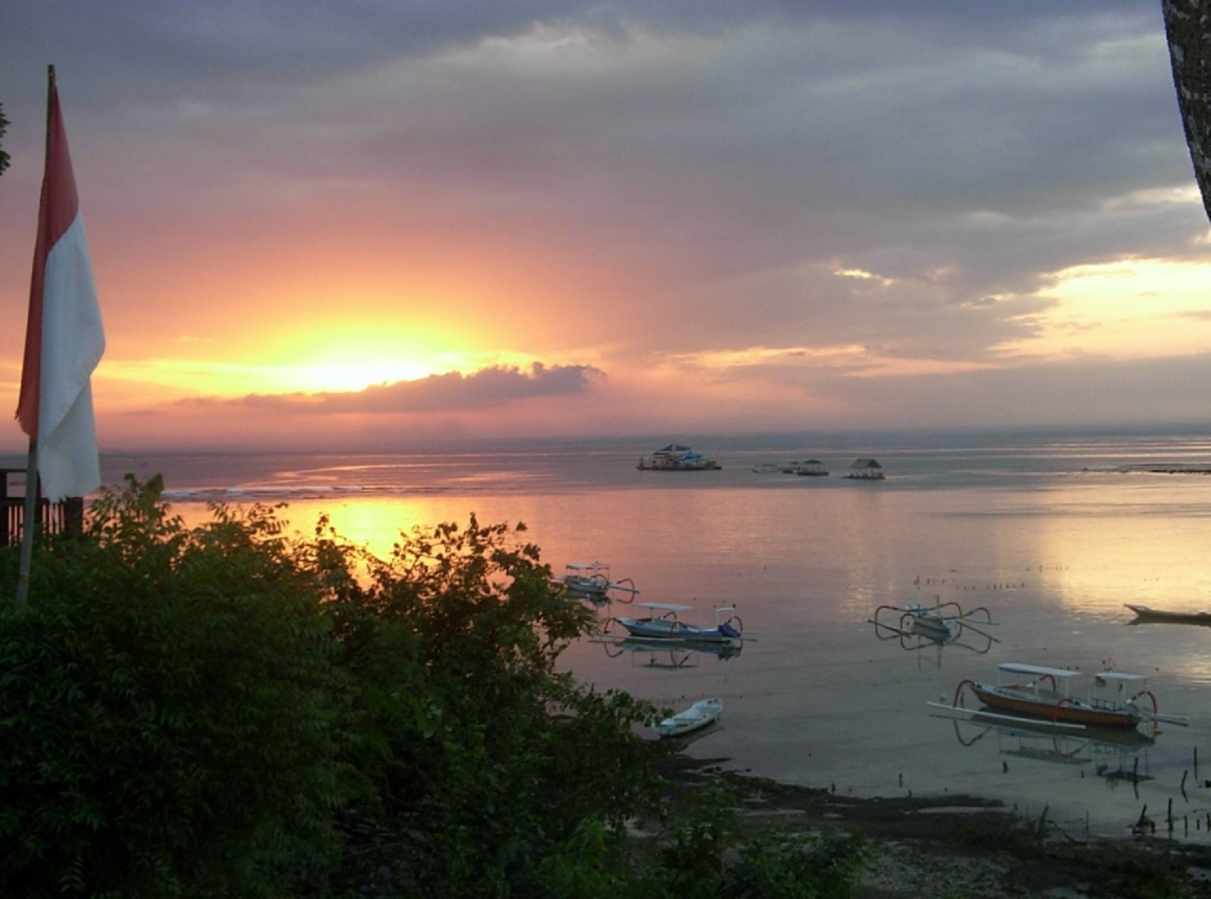 A sunset over a calm sea with several boats and a flag in the foreground.