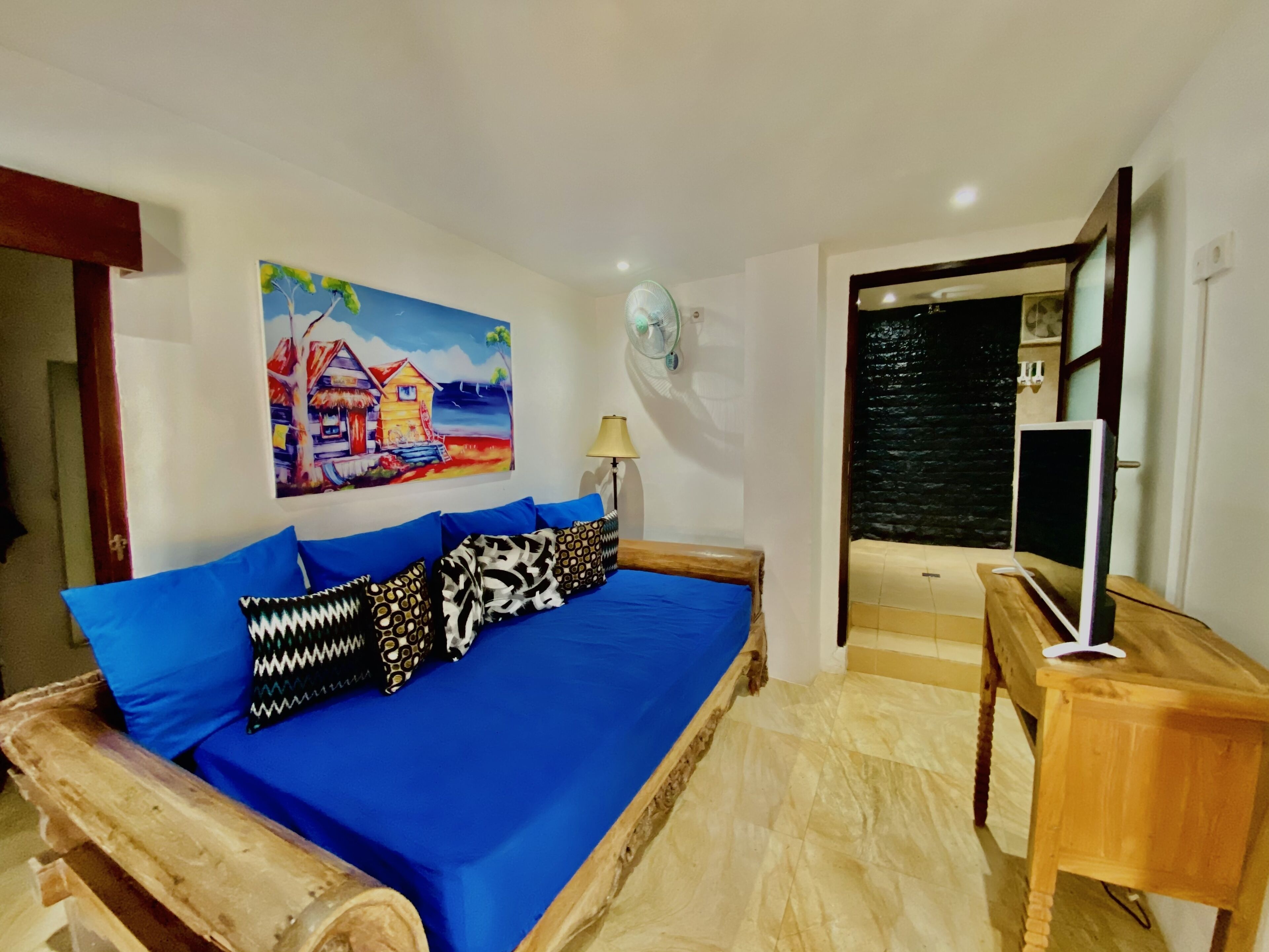 A bedroom with a bed, a painting, a fan, and a TV.