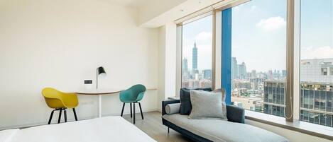A modern hotel room with a large bed, a desk, two chairs, a sofa, and a city view through large windows.