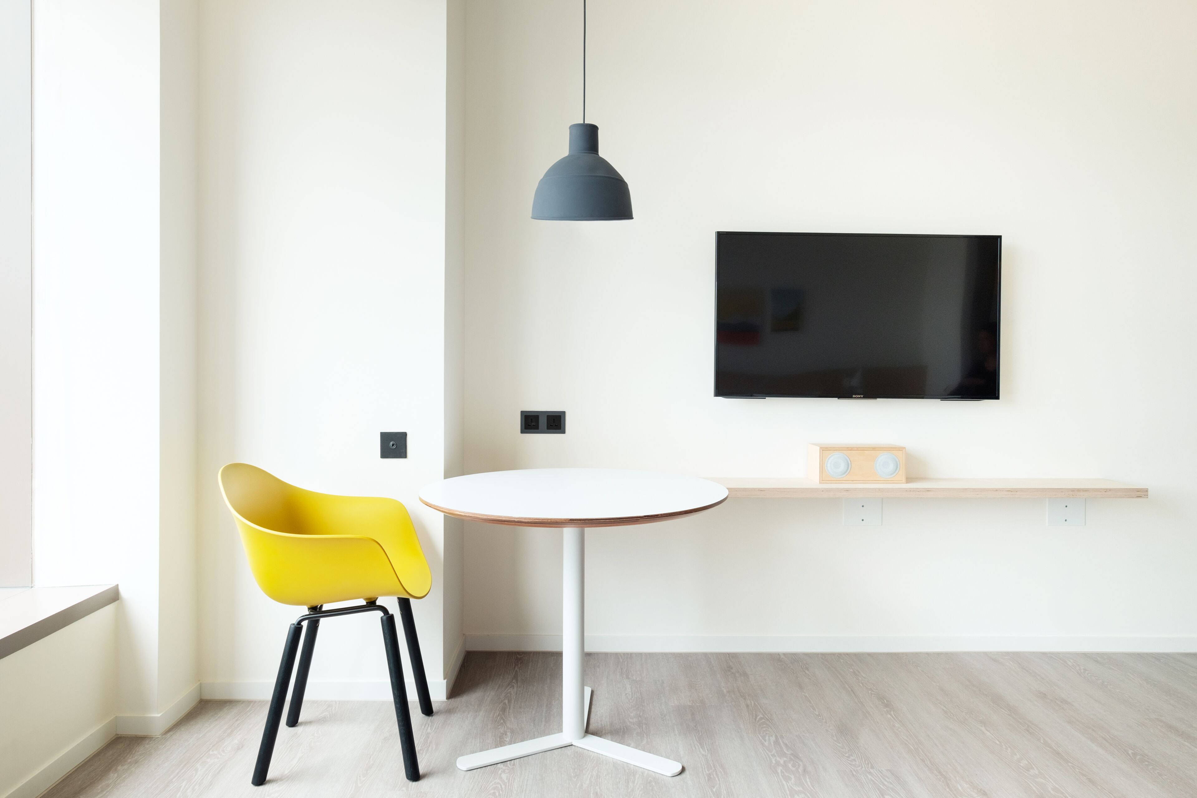 A modern room with a white wall, a round white table, a yellow chair, a black pendant light, and a flat-screen TV mounted above a wooden shelf.