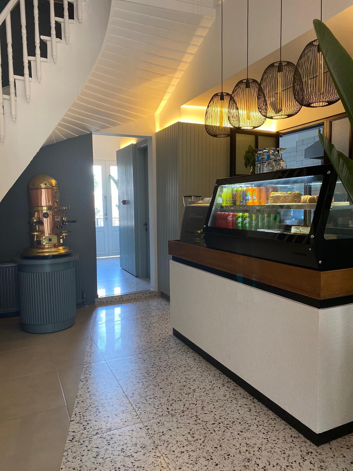 A modern interior with a counter displaying beverages and snacks, a unique cylindrical sculpture, and a staircase with white railings.