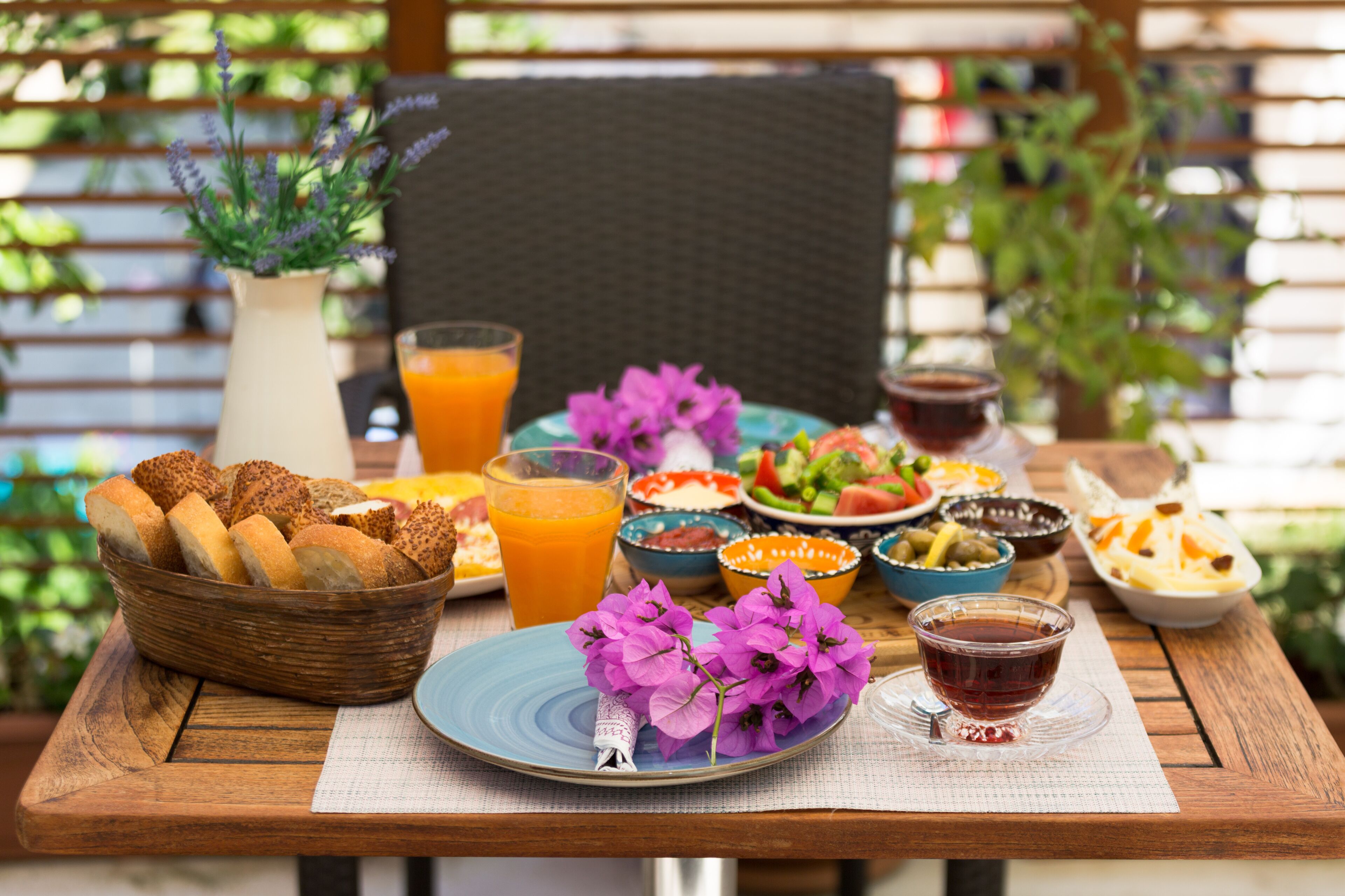 A wooden table with a variety of dishes, including bread, salad, and drinks, set outdoors with a chair and plants in the background.