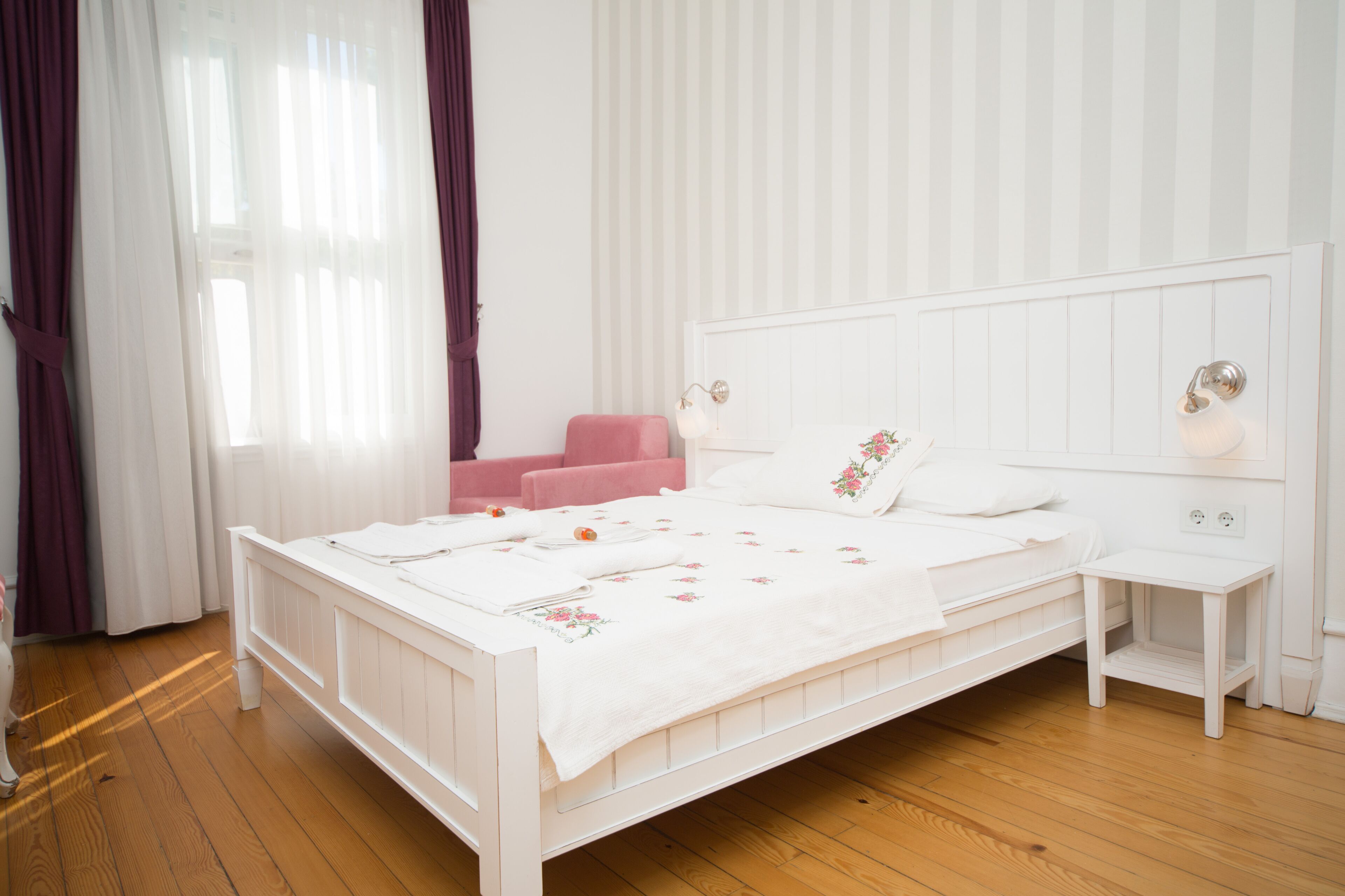 A white bed with a headboard, a bedside table, and a pink chair in a room with wooden flooring and vertical striped wallpaper.