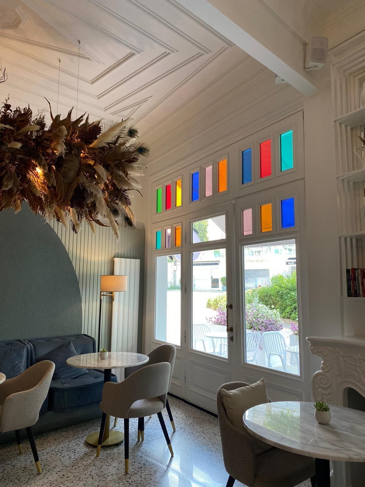 A room with a blue sofa, two round tables, and chairs. There is a large window with colorful panes and a view of the outside.