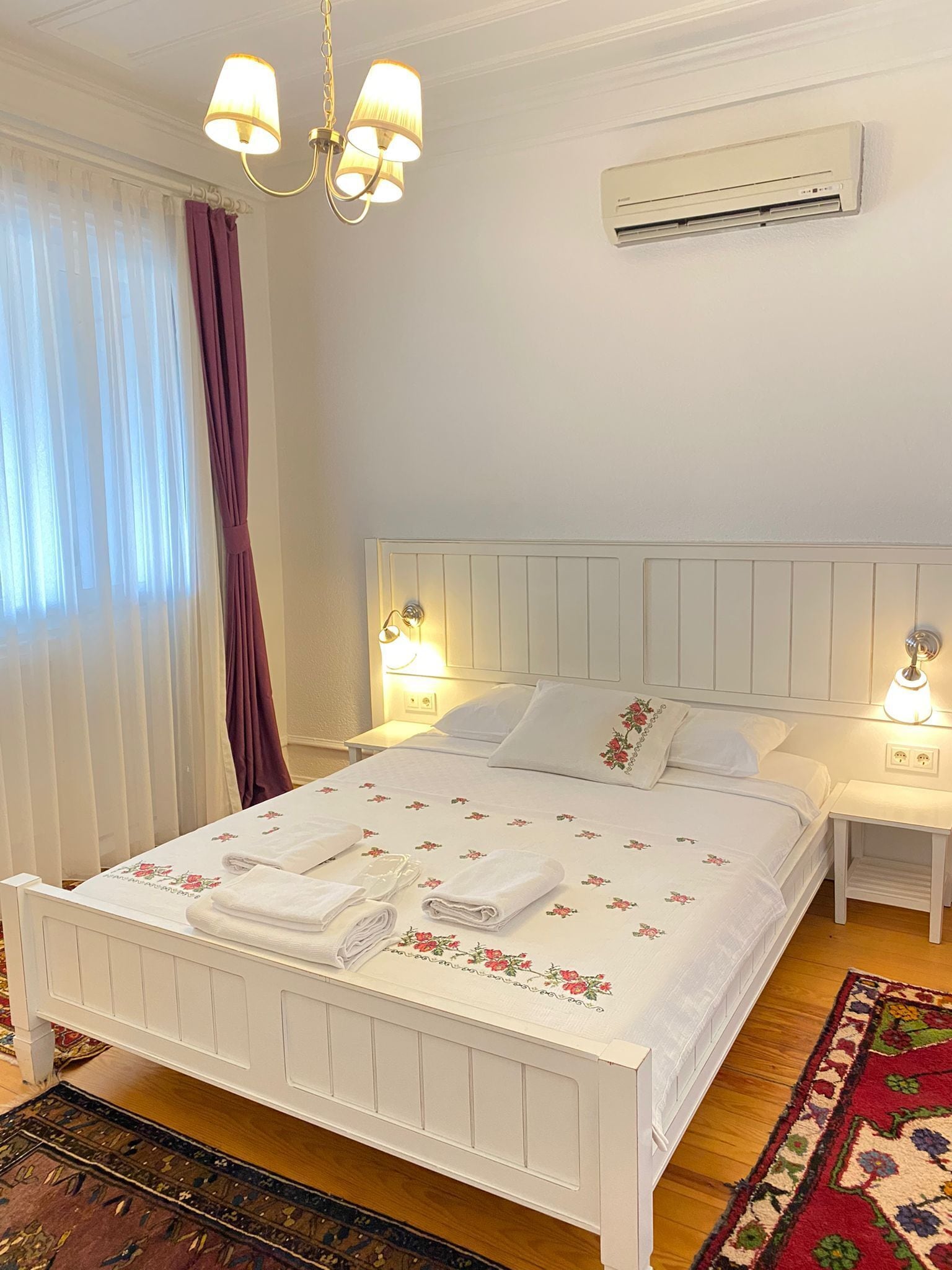 Un lit bien fait, avec du linge de lit blanc orné de motifs floraux, deux tables de chevet avec des lampes, un luminaire au plafond et un appareil de climatisation fixé au mur.