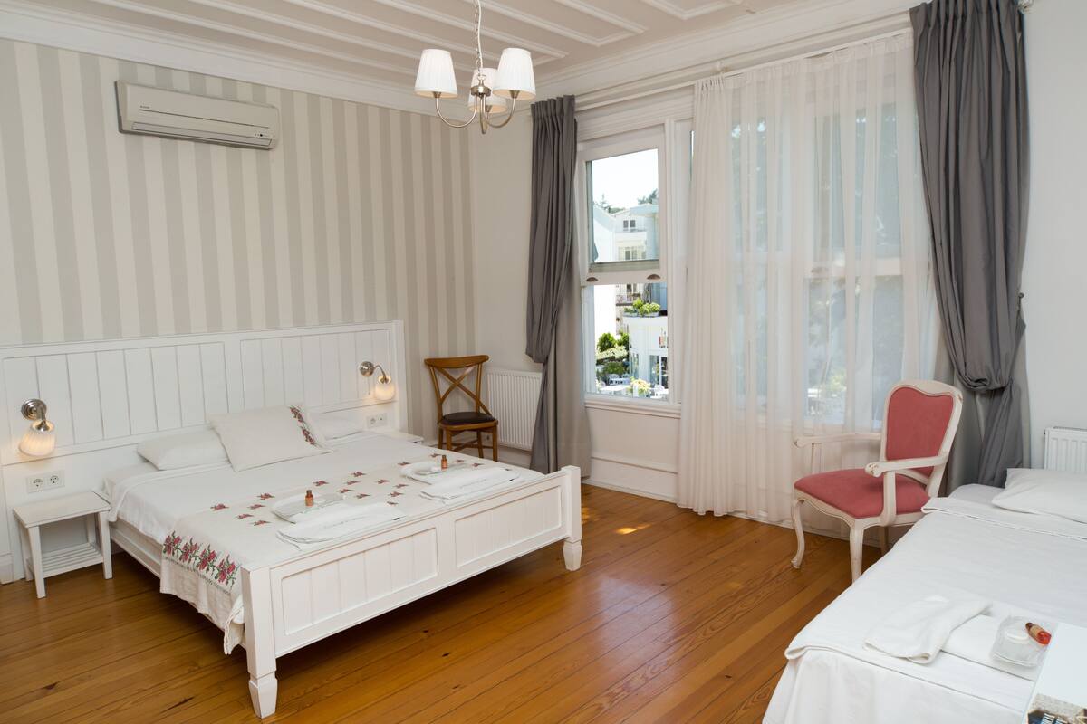 Un dormitorio con piso de madera, una cama blanca con colcha estampada, una silla de madera y un ventanal con cortinas.