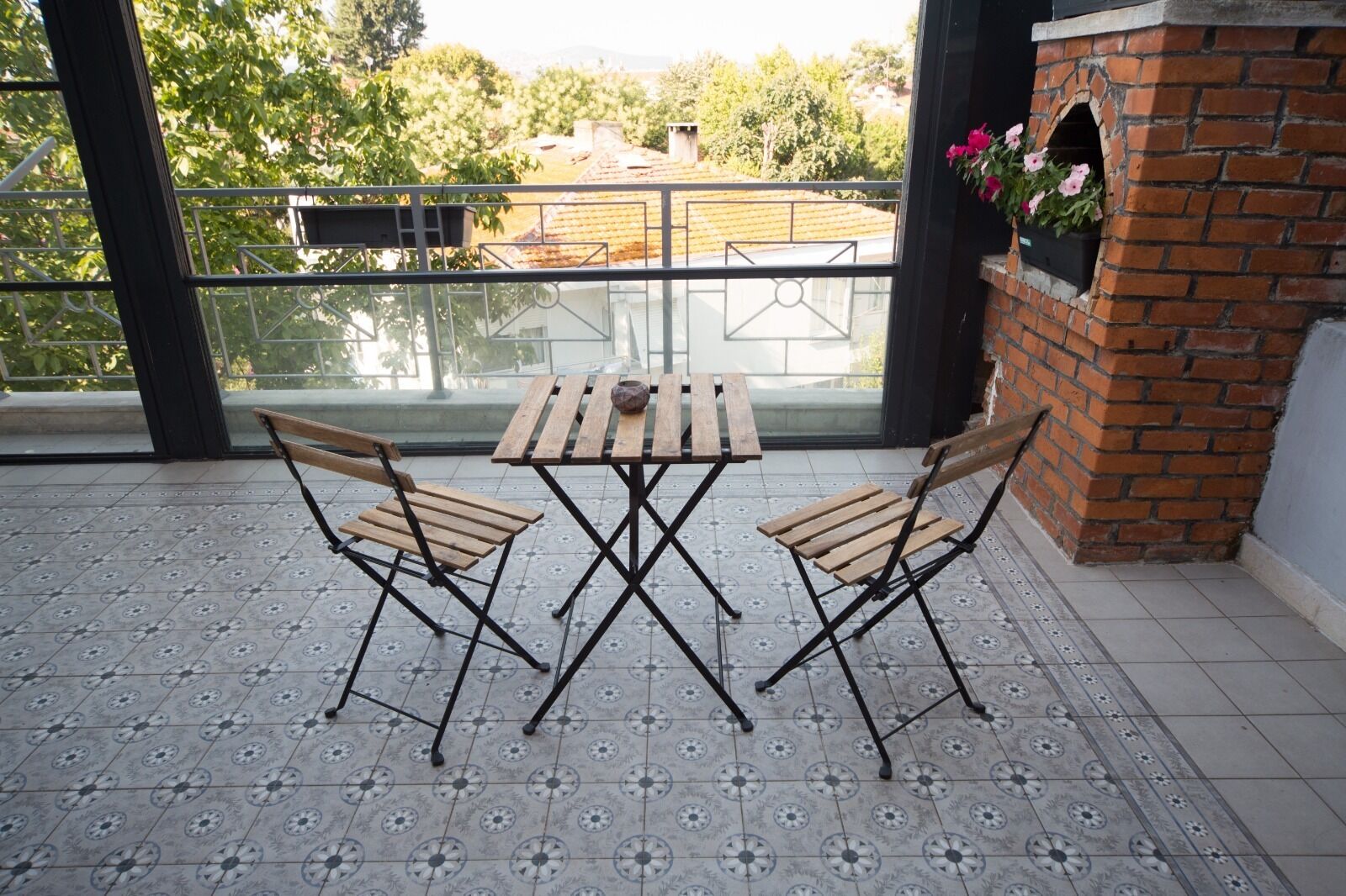 Ein Balkon mit Fliesenboden, zwei Holzstühle, ein Holztisch und ein Backofen aus Ziegelsteinen mit Blumen.