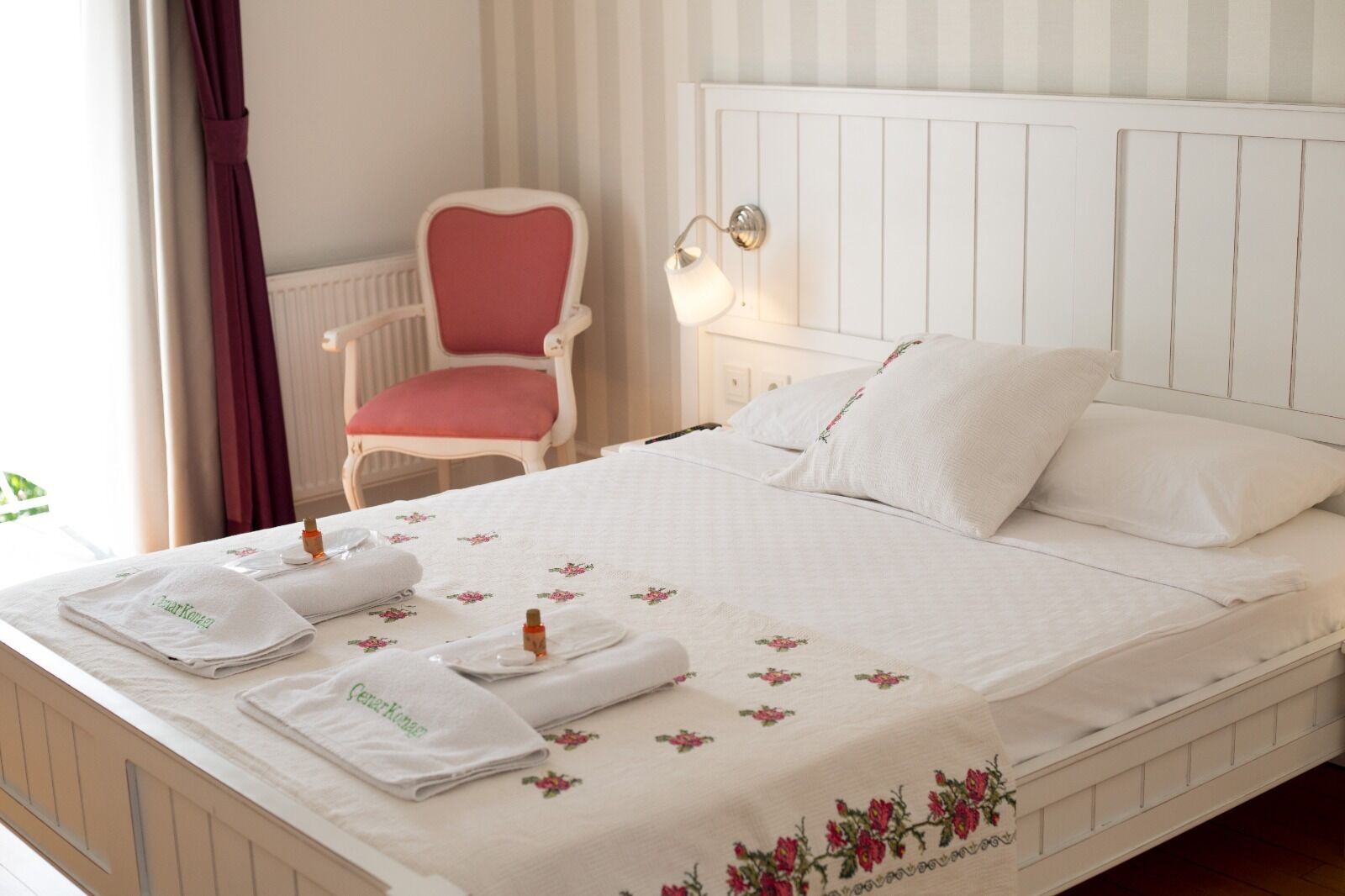 Una cama bien hecha con sábanas blancas y motivos florales, una mesita de noche con una lámpara, una silla con un cojín rojo y una ventana con cortinas.