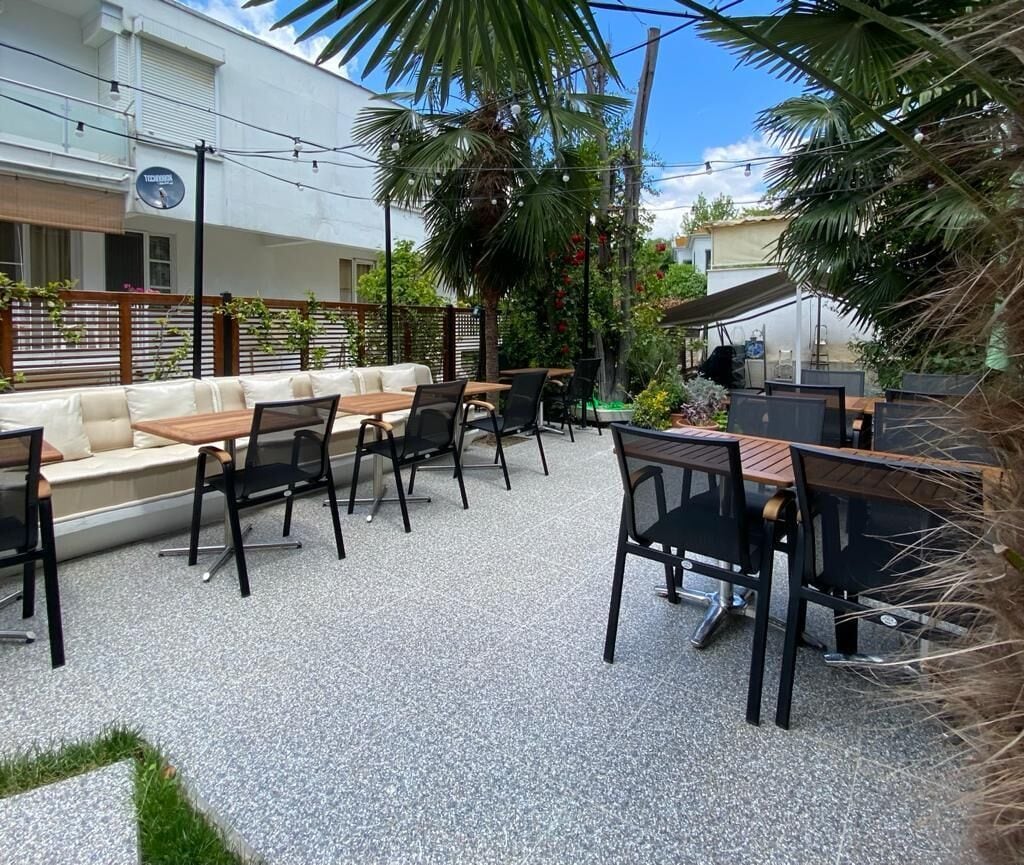 An outdoor patio area with a long wooden bench, several tables and chairs, and a small grill.