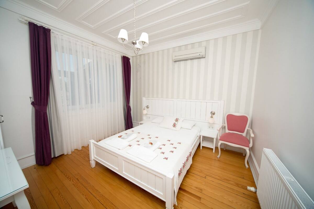 Un dormitorio con una cama blanca, una mesita de noche, una silla, un radiador y una ventana con cortinas.