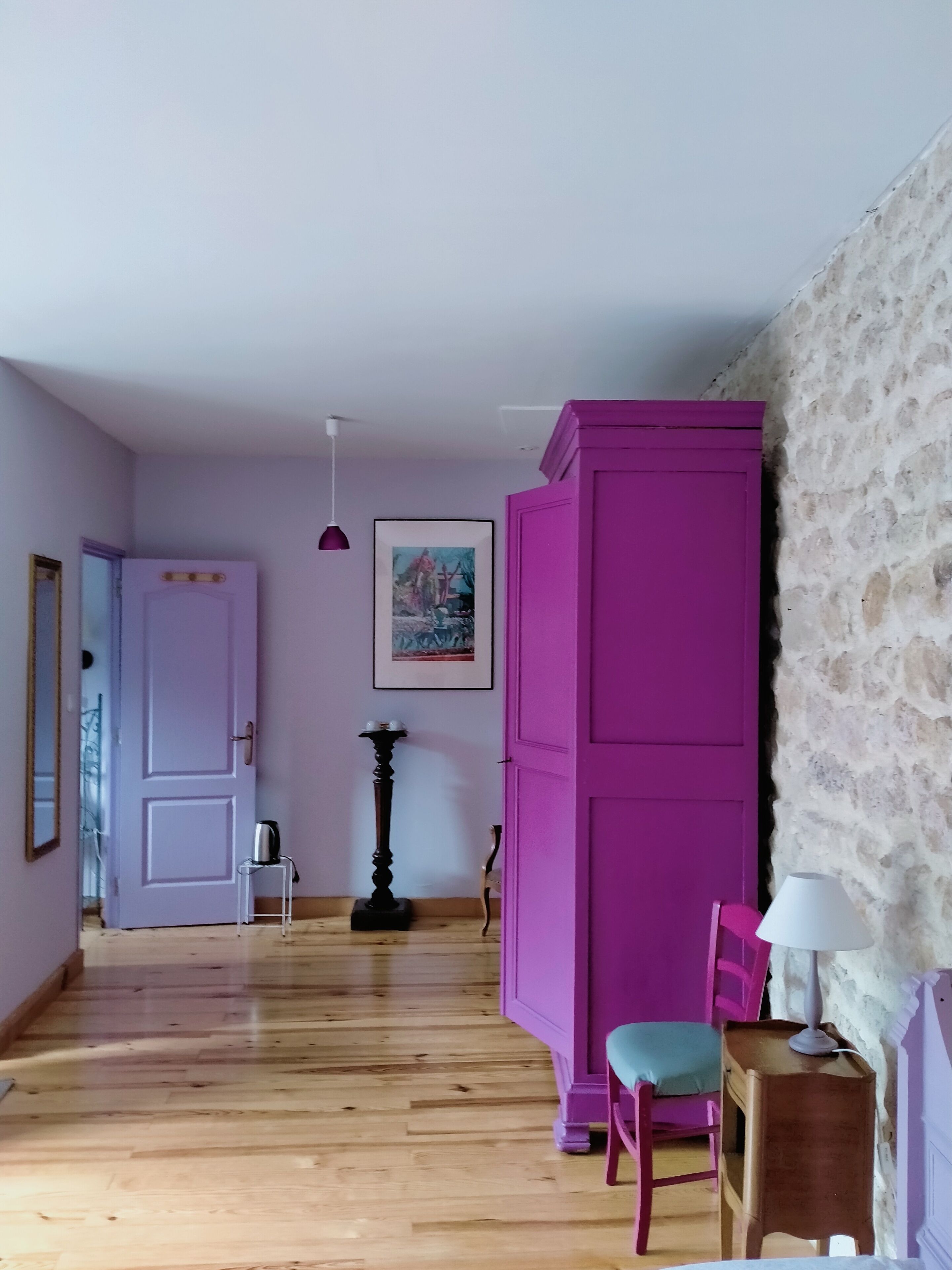 A room with wooden flooring, a purple wardrobe, a chair, a table with a lamp, and a painting on the wall.