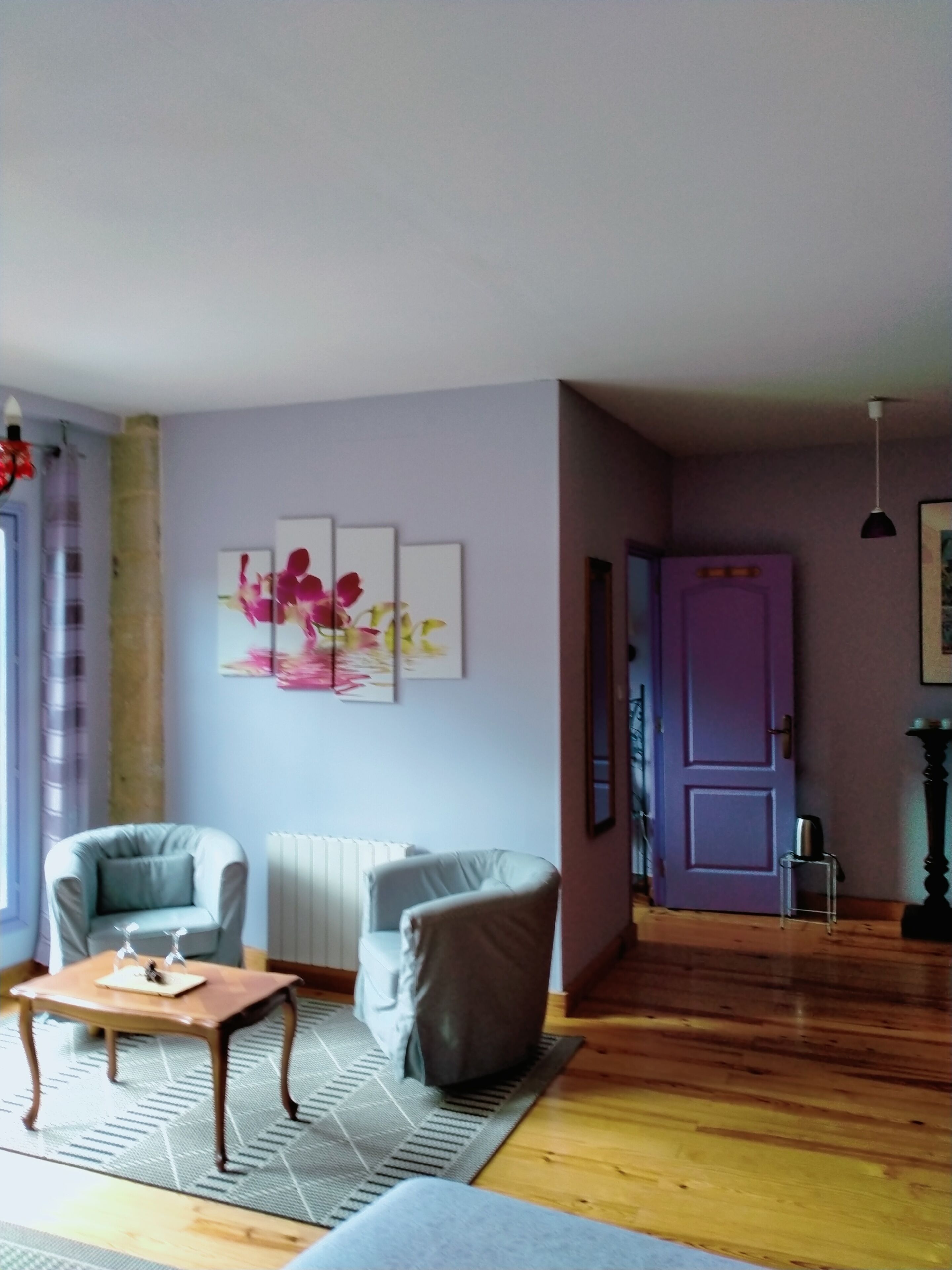 A living room with two armchairs, a wooden coffee table, and a radiator. There is a purple door and a framed picture on the wall.