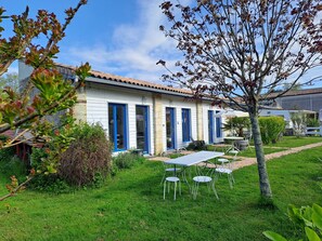 Un edificio blanco con puertas y ventanas azules, un patio con sillas y mesa de metal, y un césped bien cuidado.