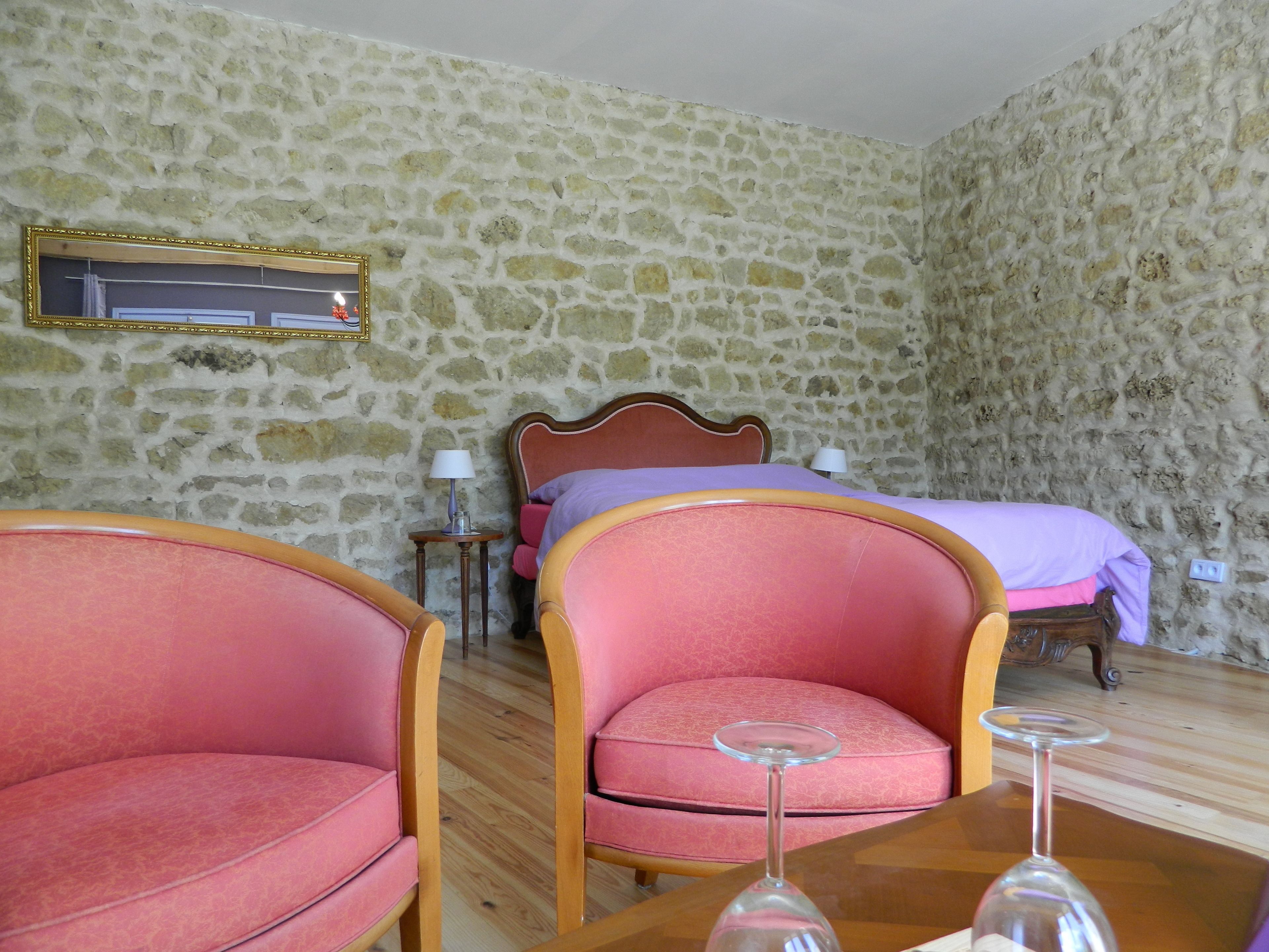 A room with a stone wall, a bed with a wooden headboard, two pink armchairs, a small table with a wine glass, and a lamp on a side table.