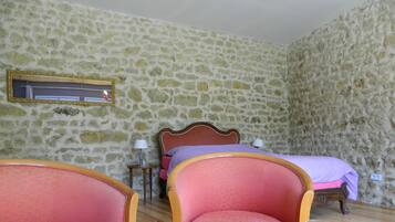 A room with a stone wall, a bed with a wooden headboard, two pink armchairs, a small table with a wine glass, and a lamp on a side table.