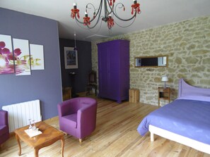Una habitación con una cama, un sillón morado, una mesa de madera, una araña y una pared de piedra.