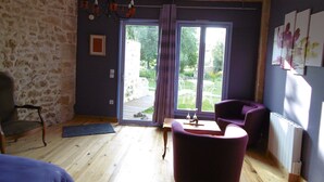 A room with a stone wall, a wooden floor, a chandelier, a purple chair, a table, and a view of a garden through a glass door. - La Cadournaise (Saint-Seurin-de-Cadourne)