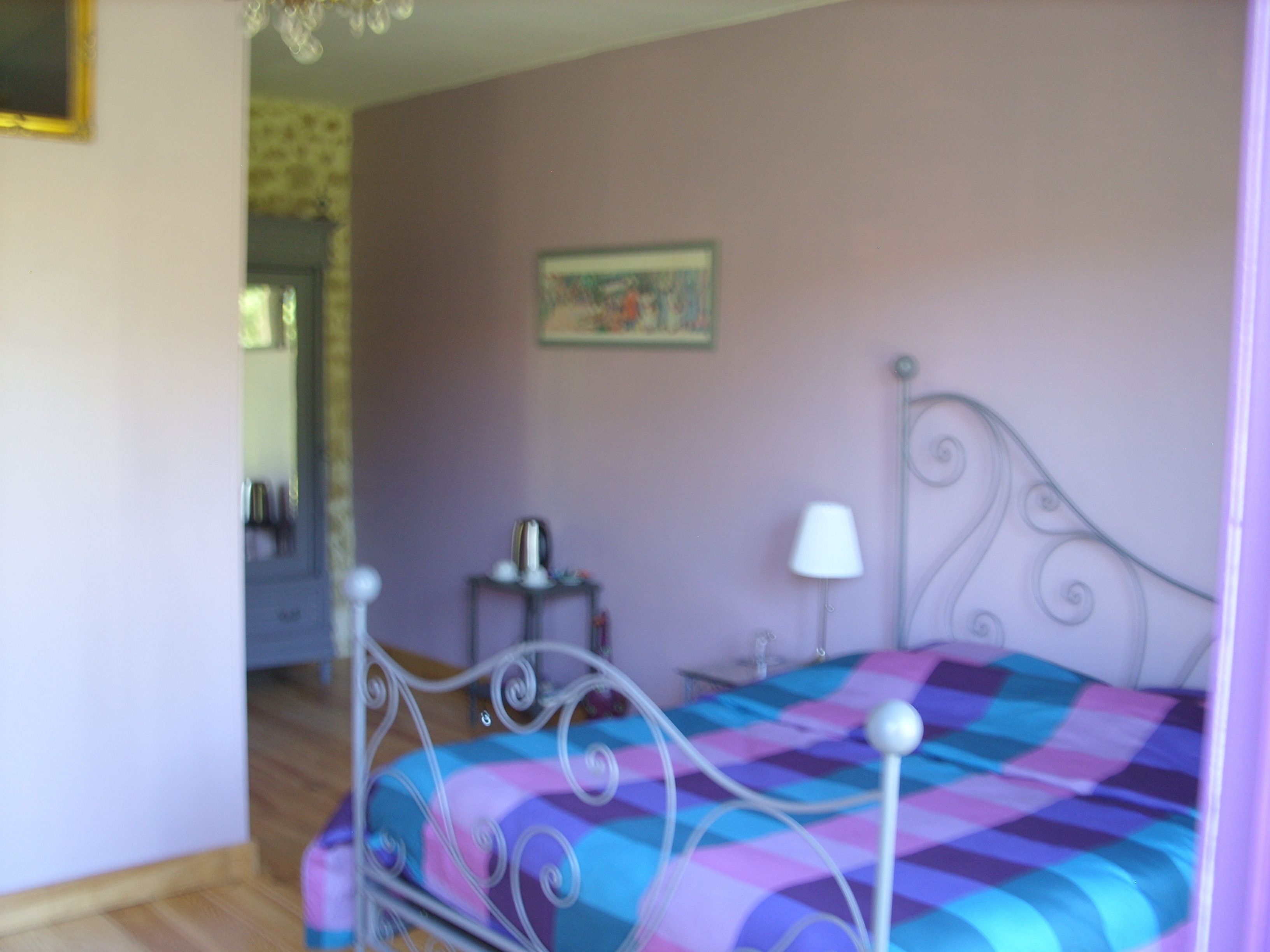 A bedroom with a metal bed frame, a colorful bedspread, a bedside table with a lamp, and a small decorative item on the wall.