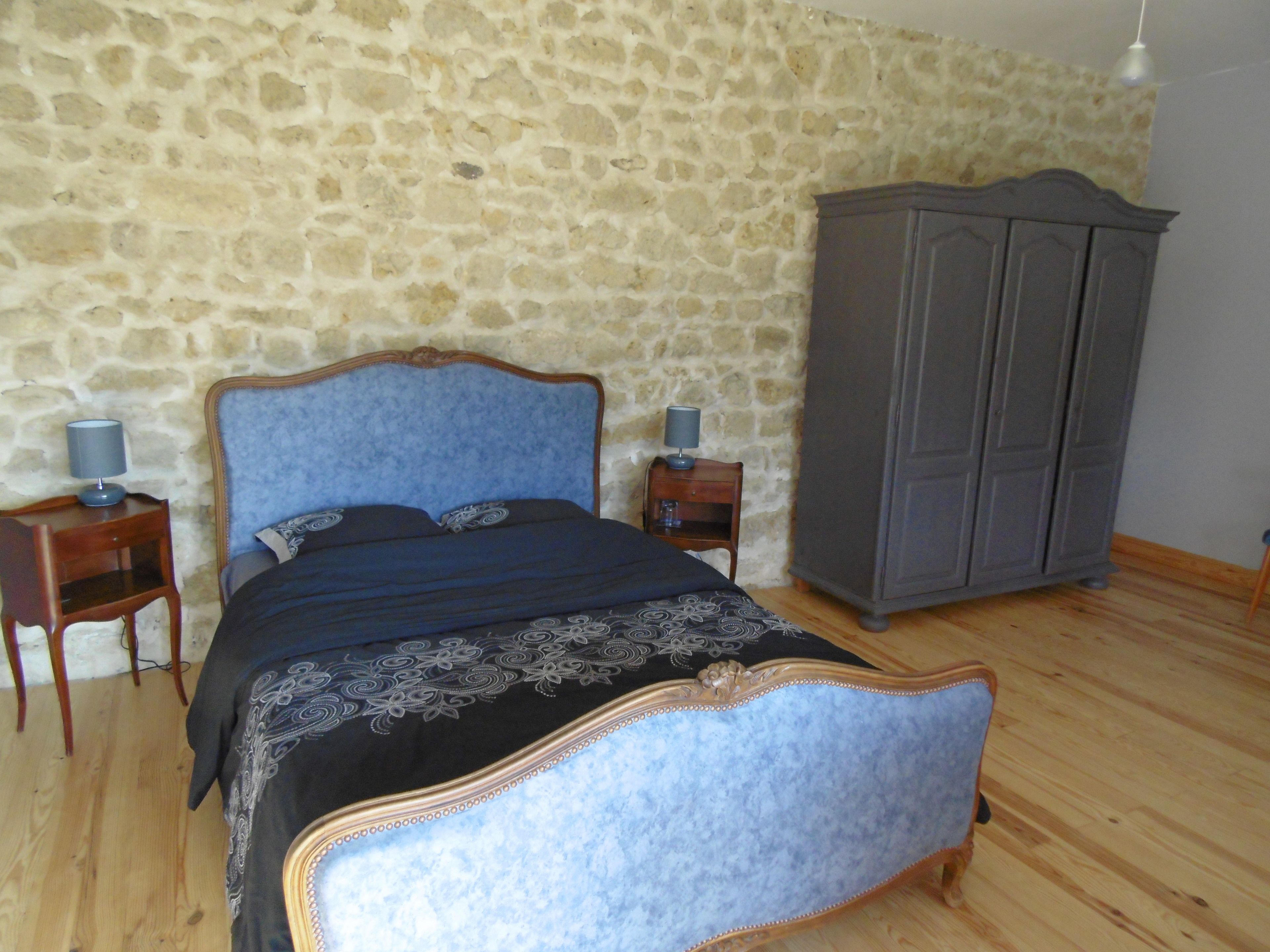 A bedroom with a stone wall, a bed with a blue headboard, two bedside tables with lamps, and a wardrobe.