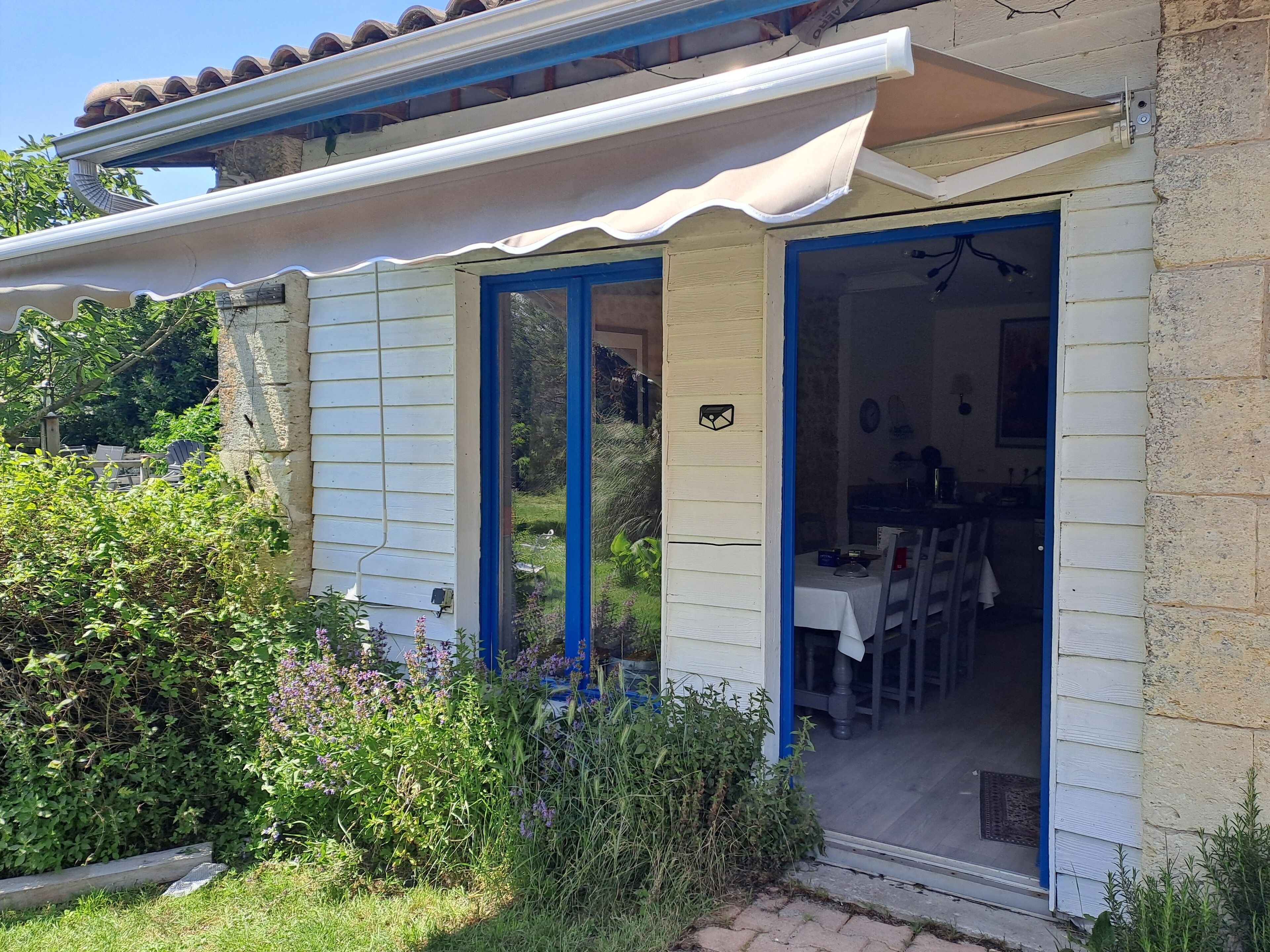 Une maison dotée d’un porche abrité, d’un coin repas visible à travers la porte ouverte et d’un jardin où poussent différentes espèces de plantes.