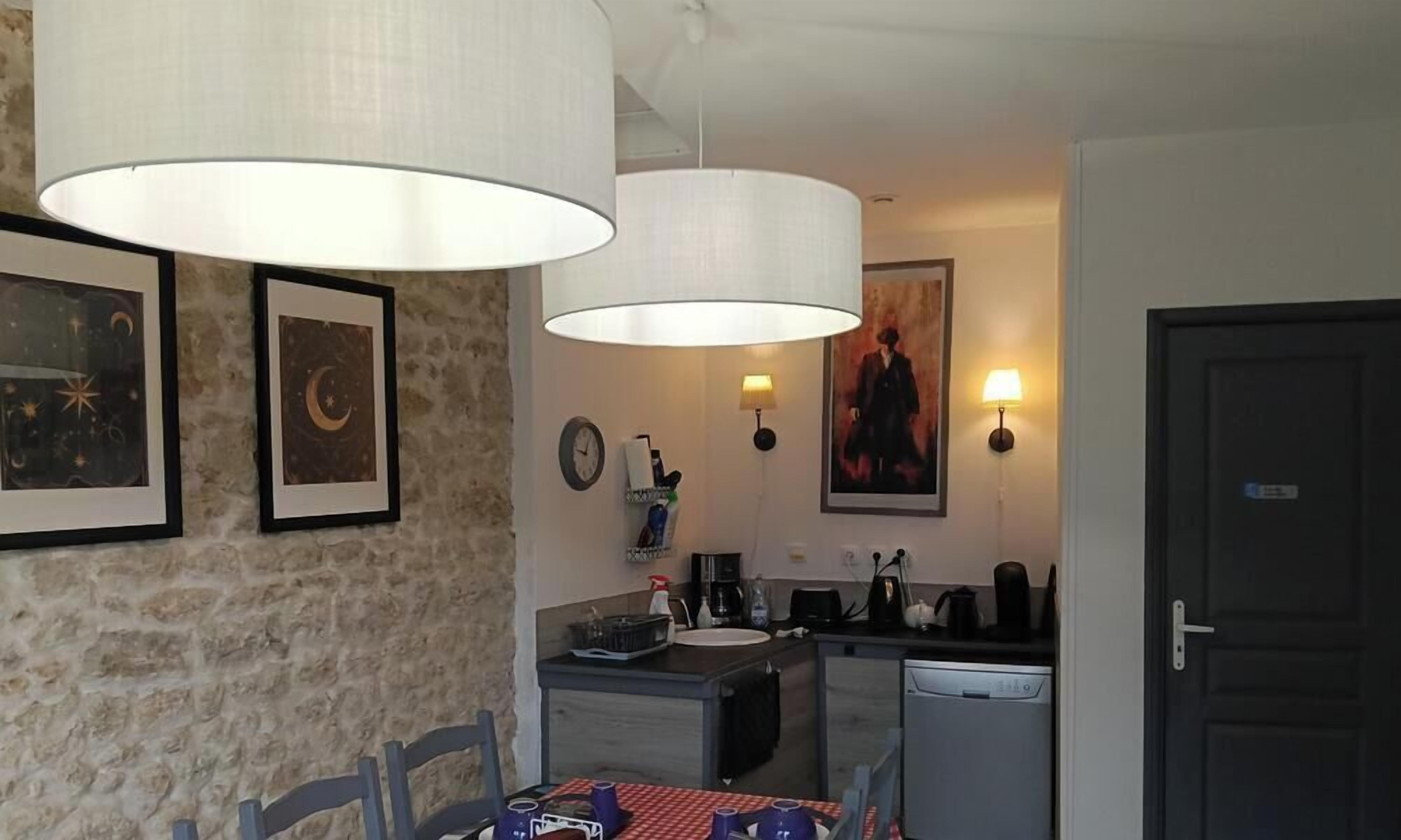 Une cuisine compacte équipée d’un évier, d’une machine à café et d’un petit réfrigérateur. Deux lampes suspendues sont fixées au-dessus du comptoir, et une horloge est accrochée au mur.