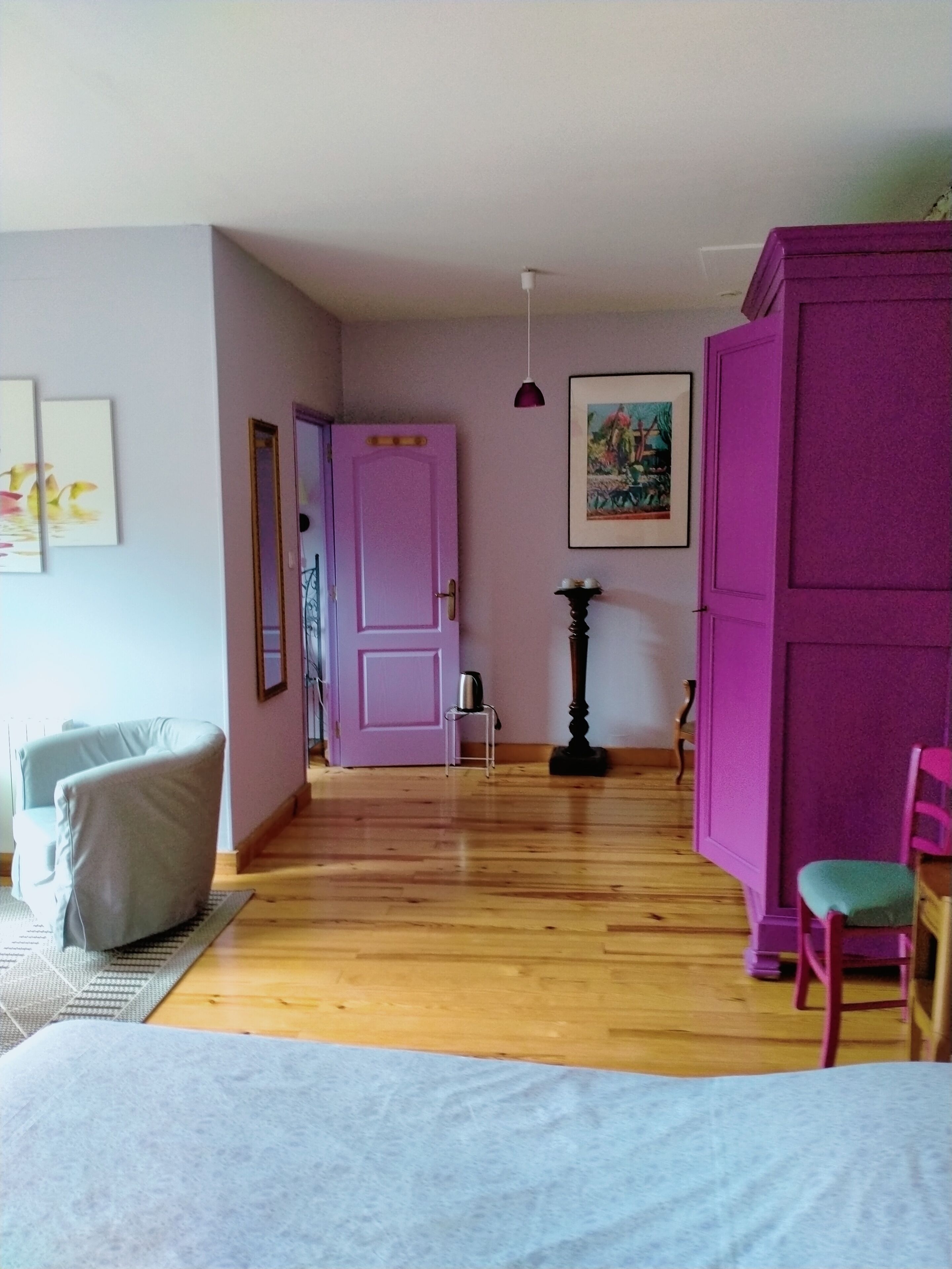 Une pièce avec un plancher en bois, un lit, une chaise, une petite table, une porte rose et un tableau accroché au mur.
