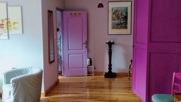 Une pièce avec un plancher en bois, un lit, une chaise, une petite table, une porte rose et un tableau accroché au mur.