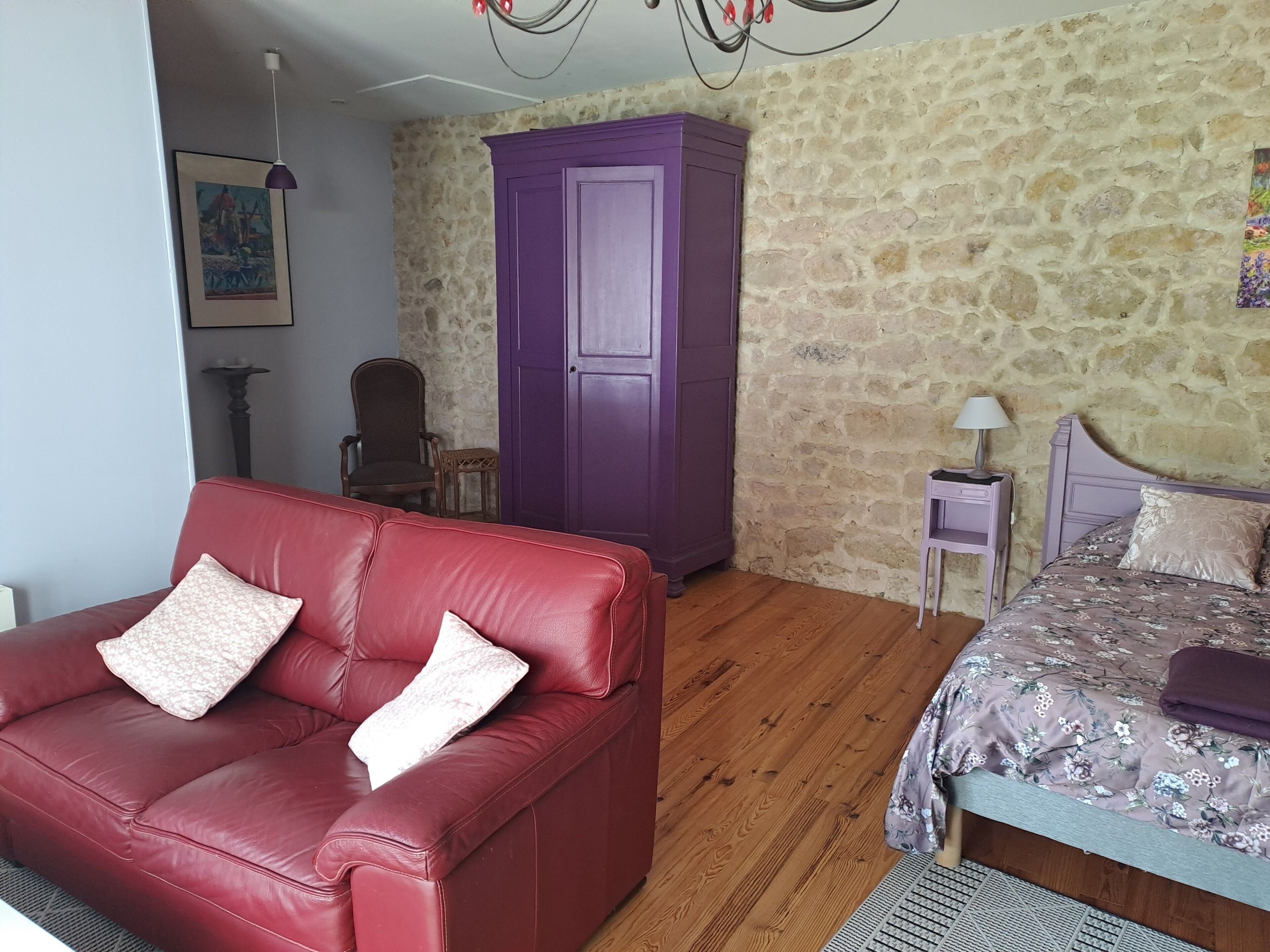 A room with a red leather sofa, a purple wardrobe, a wooden floor, a bed with floral bedding, a small table with a lamp, and a stone wall.