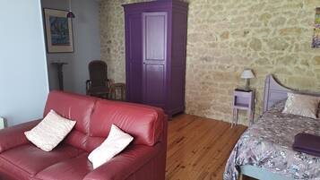 A room with a red leather sofa, a purple wardrobe, a wooden floor, a bed with floral bedding, a small table with a lamp, and a stone wall.