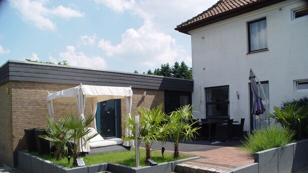 Un patio exterior moderno con un espacio de estar techado, macetas con palmeras y un sendero de ladrillo que lleva a un edificio con techo de tejas.