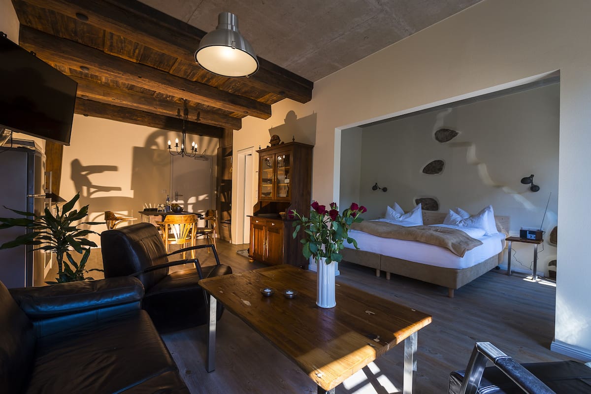 A modern living room with a wooden ceiling, a sofa, a dining table, and a bed with a white headboard.