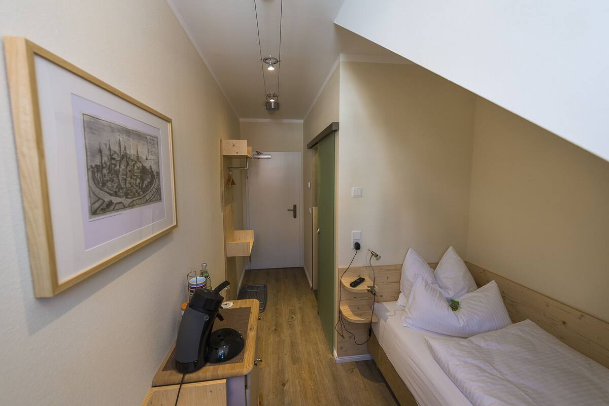 A small, narrow room with a bed, a desk with a computer, and a framed picture on the wall.