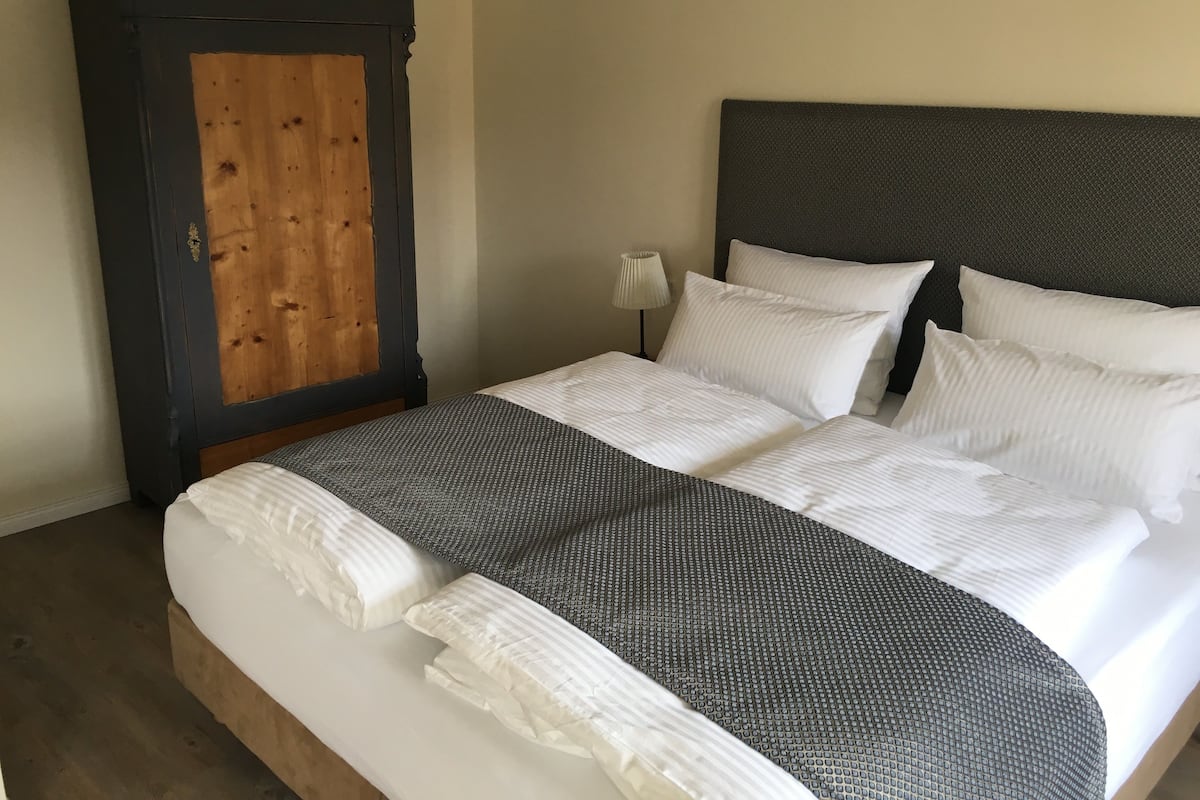 A neatly made bed with white linens and a grey patterned bedspread, a wooden wardrobe with a black frame, and a bedside lamp.