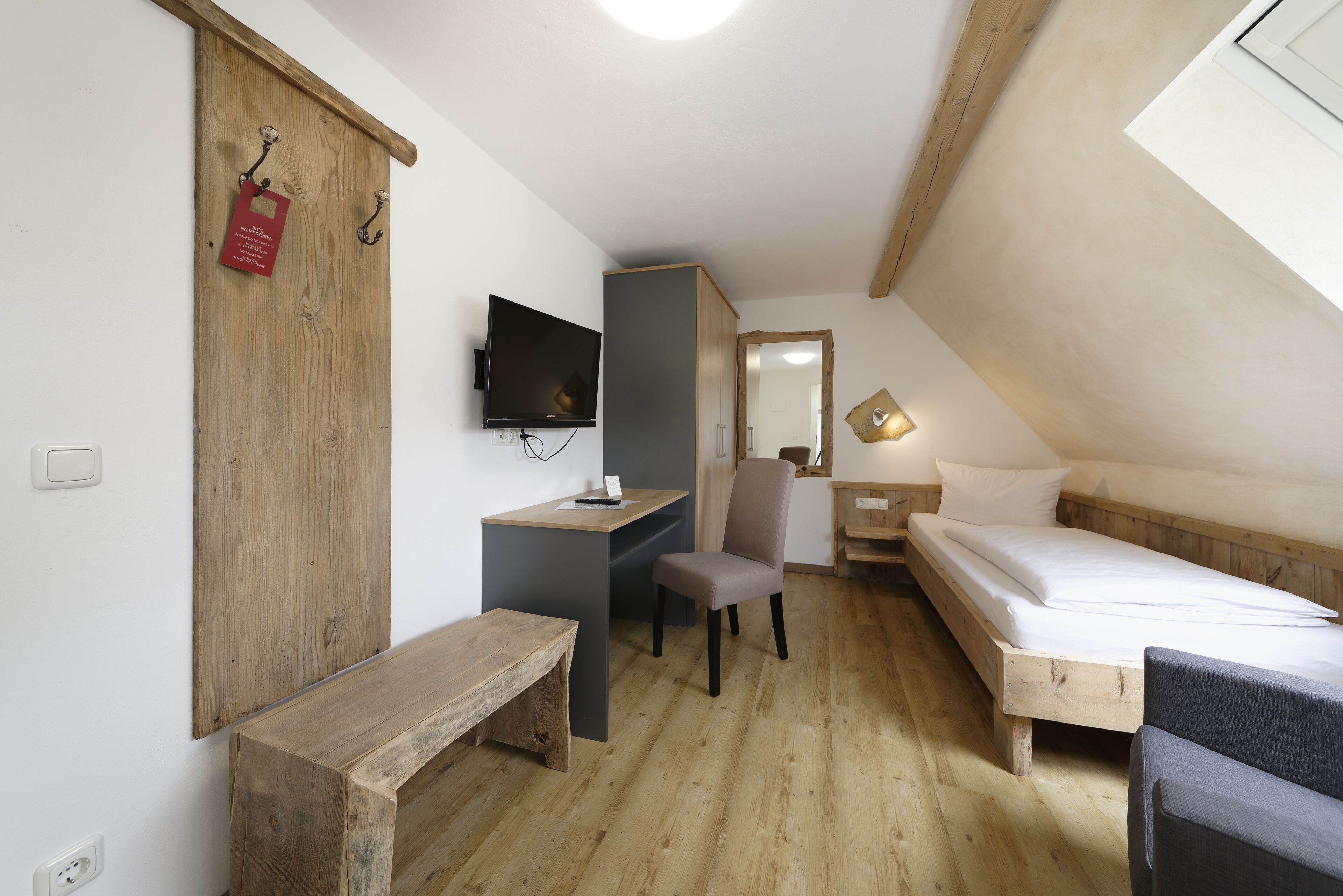 Ein kleiner, gemütlicher Raum mit einem Holztisch, einem Sessel, einem Bett und einem an der Wand befestigten Fernseher.