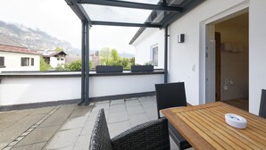 Un balcón con una mesa y sillas de madera, una puerta de cristal que da al baño, y una vista de casas y montañas al fondo.