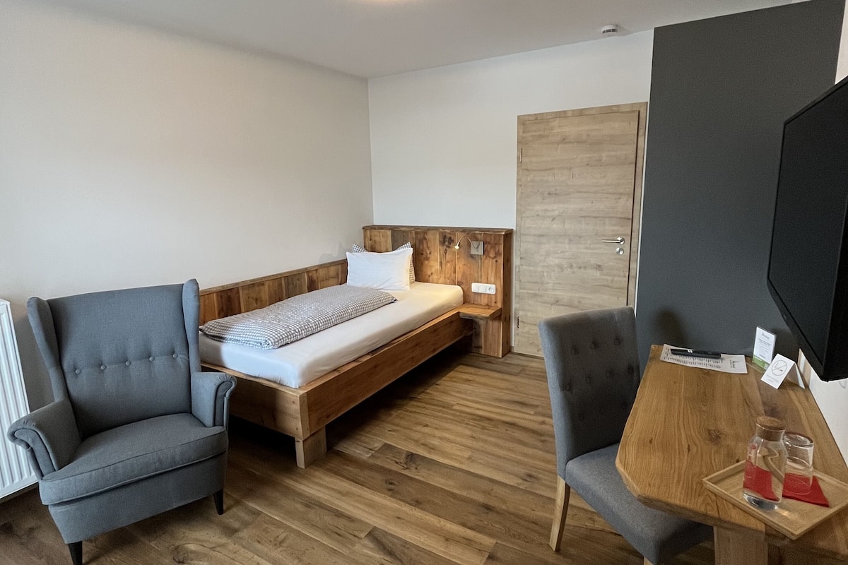 A modern hotel room with a wooden floor, a bed with a wooden headboard, a grey armchair, a wooden table with a tray, and a television on the right.