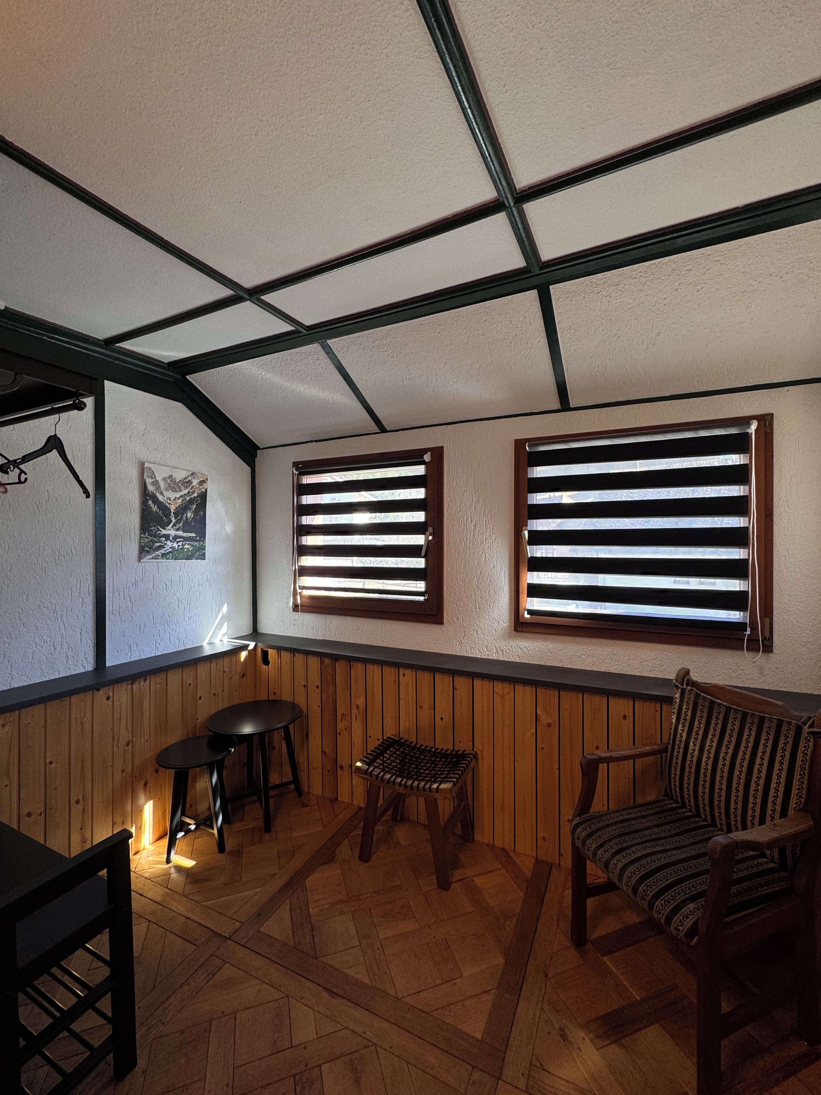 Um quarto com painéis de madeira, duas janelas com persianas, um banco de madeira, uma mesa pequena e um cabideiro.