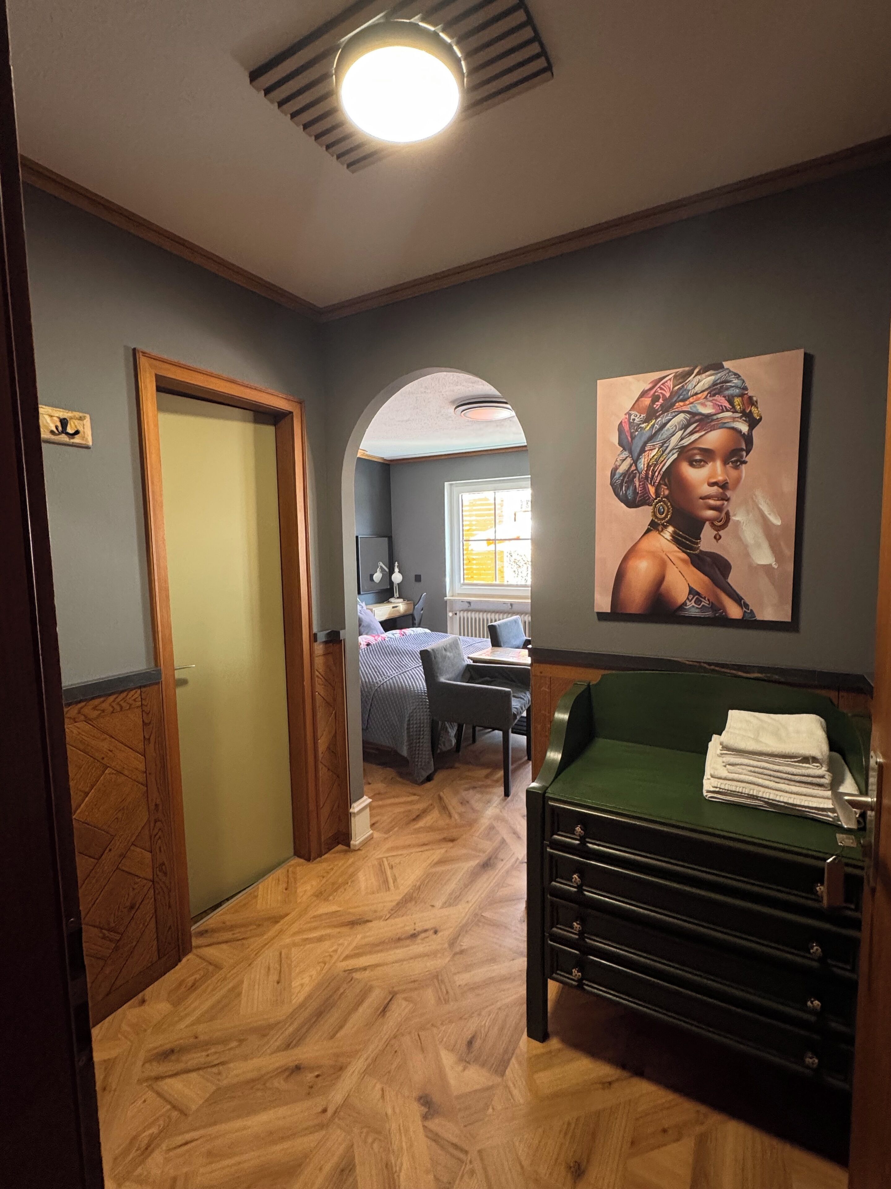 A hotel room with a wooden herringbone floor, a green sofa, a bed with a blue and white patterned bedspread, a painting on the wall, and a window with daylight coming through.