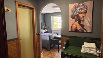 A hotel room with a wooden herringbone floor, a green sofa, a bed with a blue and white patterned bedspread, a painting on the wall, and a window with daylight coming through.