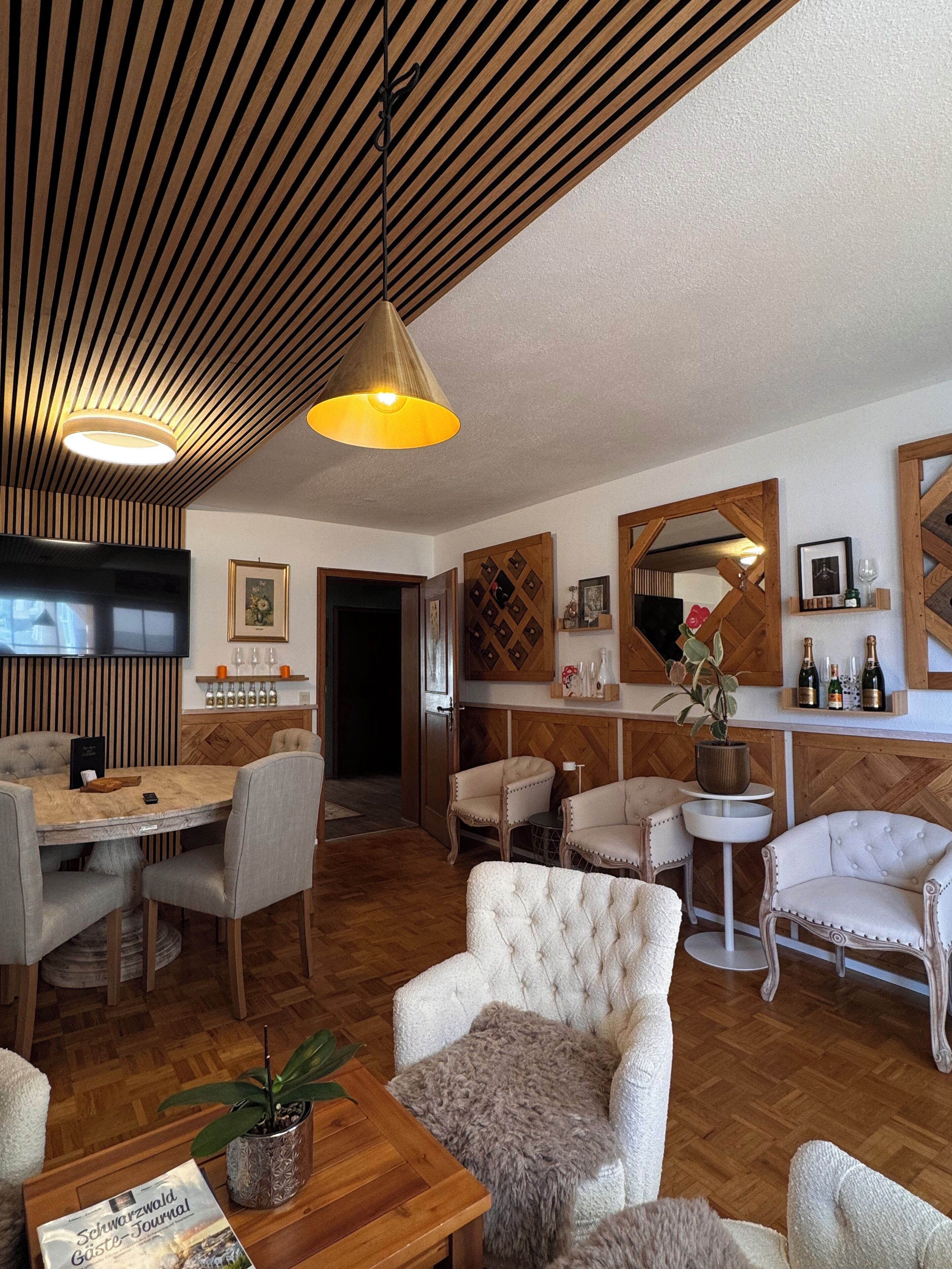 A modern living room with a wooden ceiling, a dining area with a round table, and a bar area with a mirror and framed pictures.