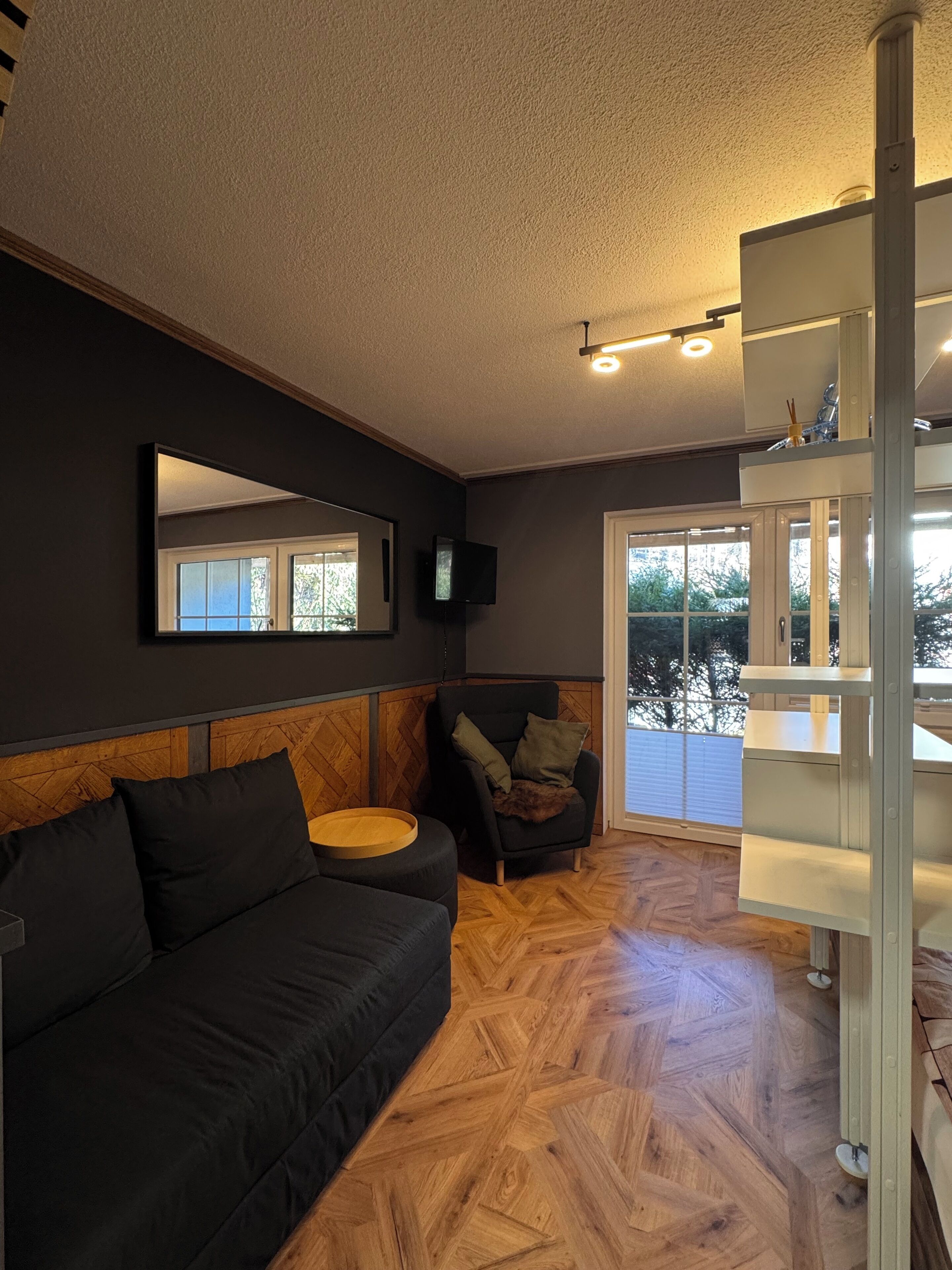 A modern living room with a black sofa, a wooden herringbone floor, a large mirror, and a television mounted on the wall.