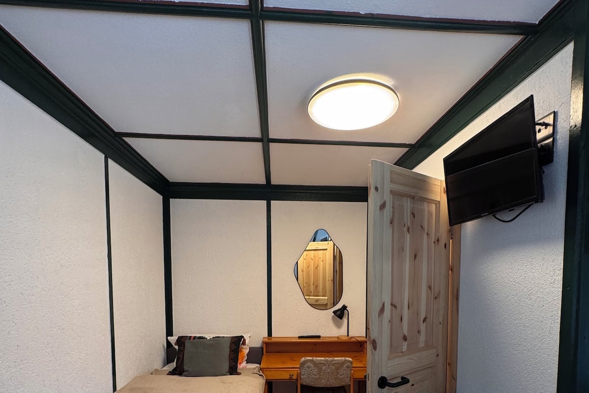 A compact room with a bed, a desk, a chair, a mirror, and a television mounted on the wall.