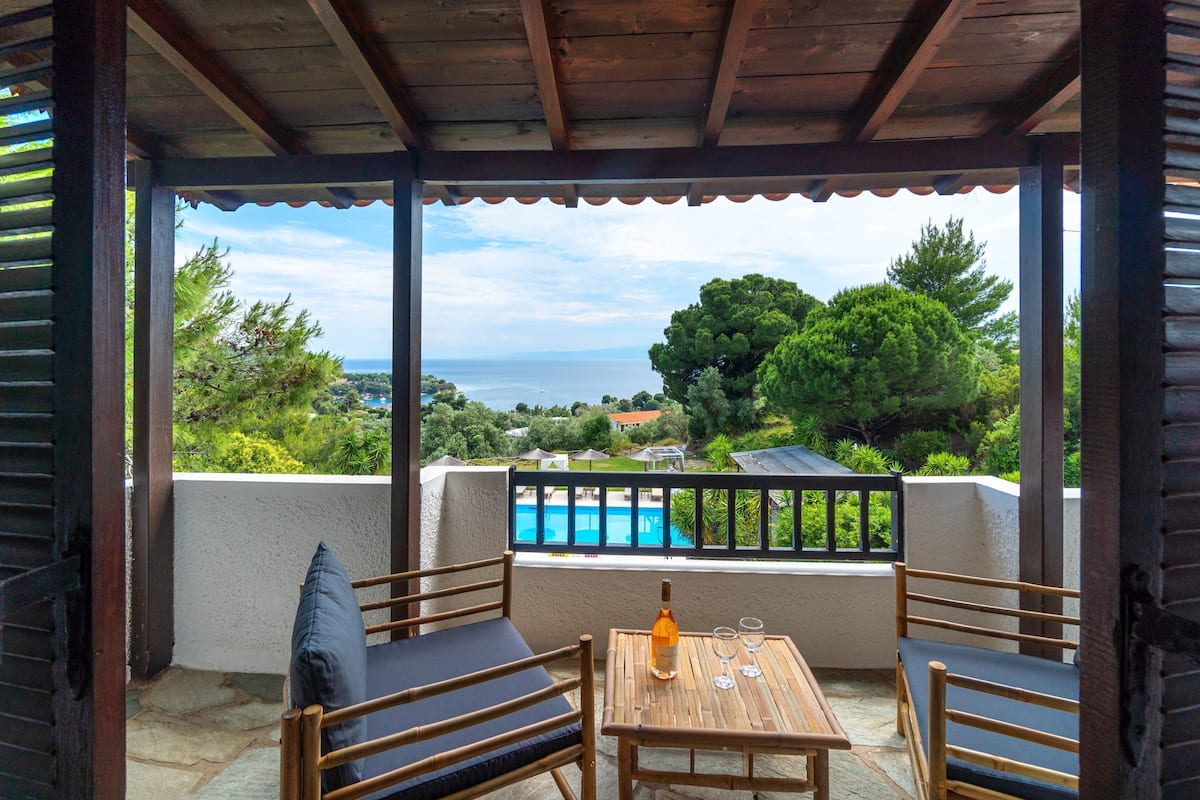 Un porche techado con una mesa de madera y dos sillas, con vista a una piscina y al mar.