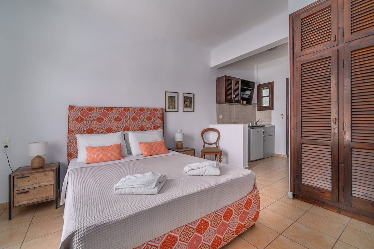 Una cama bien tendida con una colcha estampada, una mesita de noche de madera con una lámpara y un armario con puertas ribeteadas.