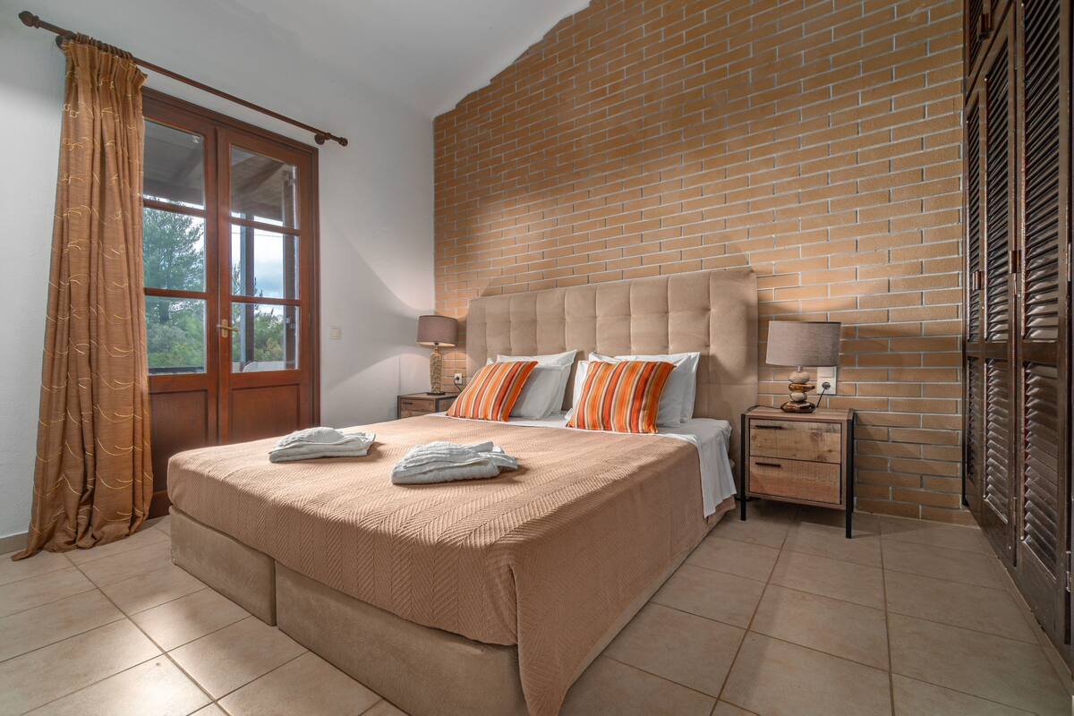 Un dormitorio con una cama grande, una pared de ladrillo, una ventana con cortinas y una mesita de noche con una lámpara.