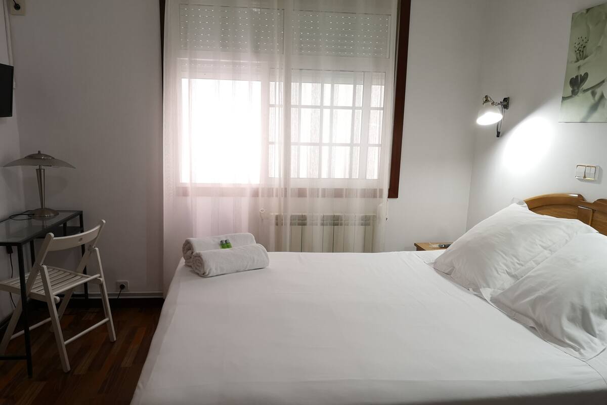 Una cama bien hecha con sábanas blancas, una silla, un escritorio con lámpara y una ventana con cortinas traslúcidas.